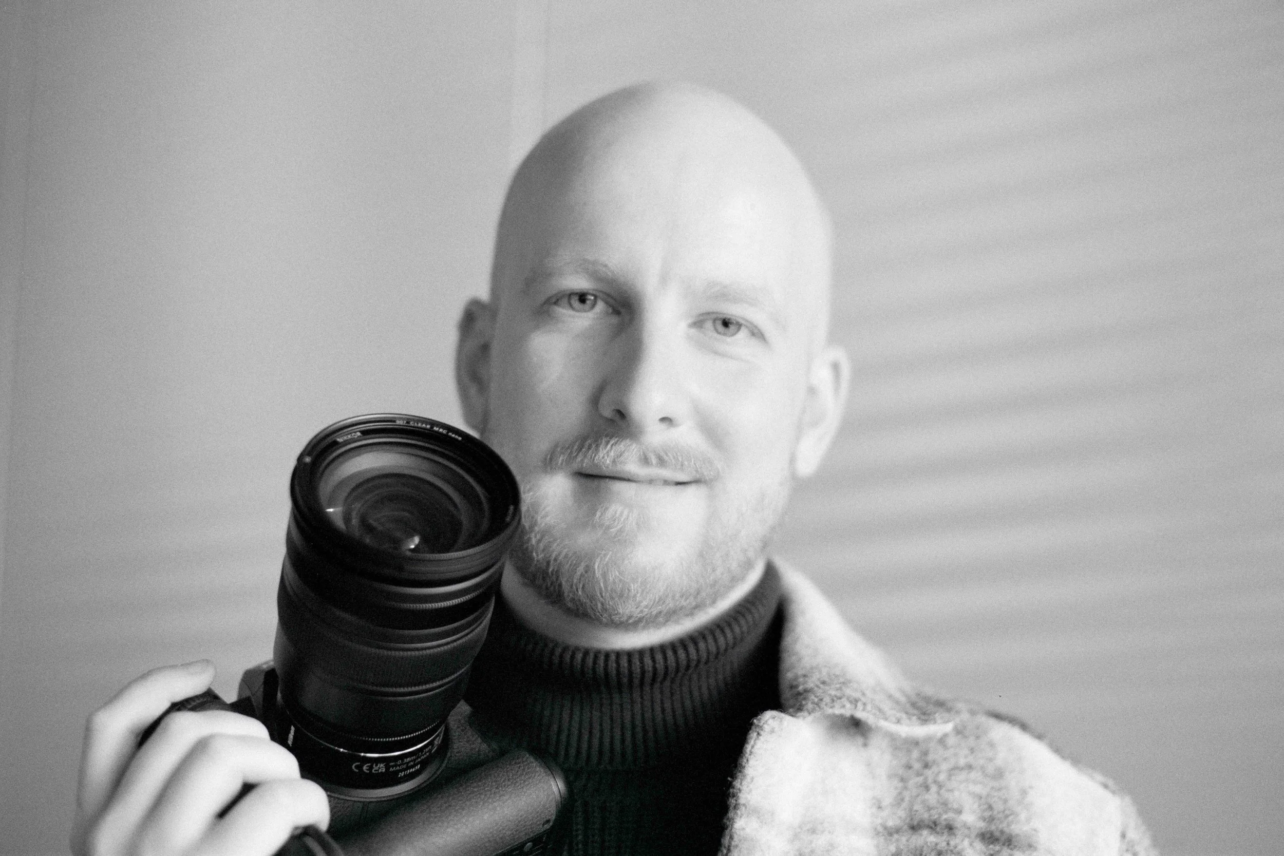 Black and white portrait of a bald man with a beard holding a camera lens.