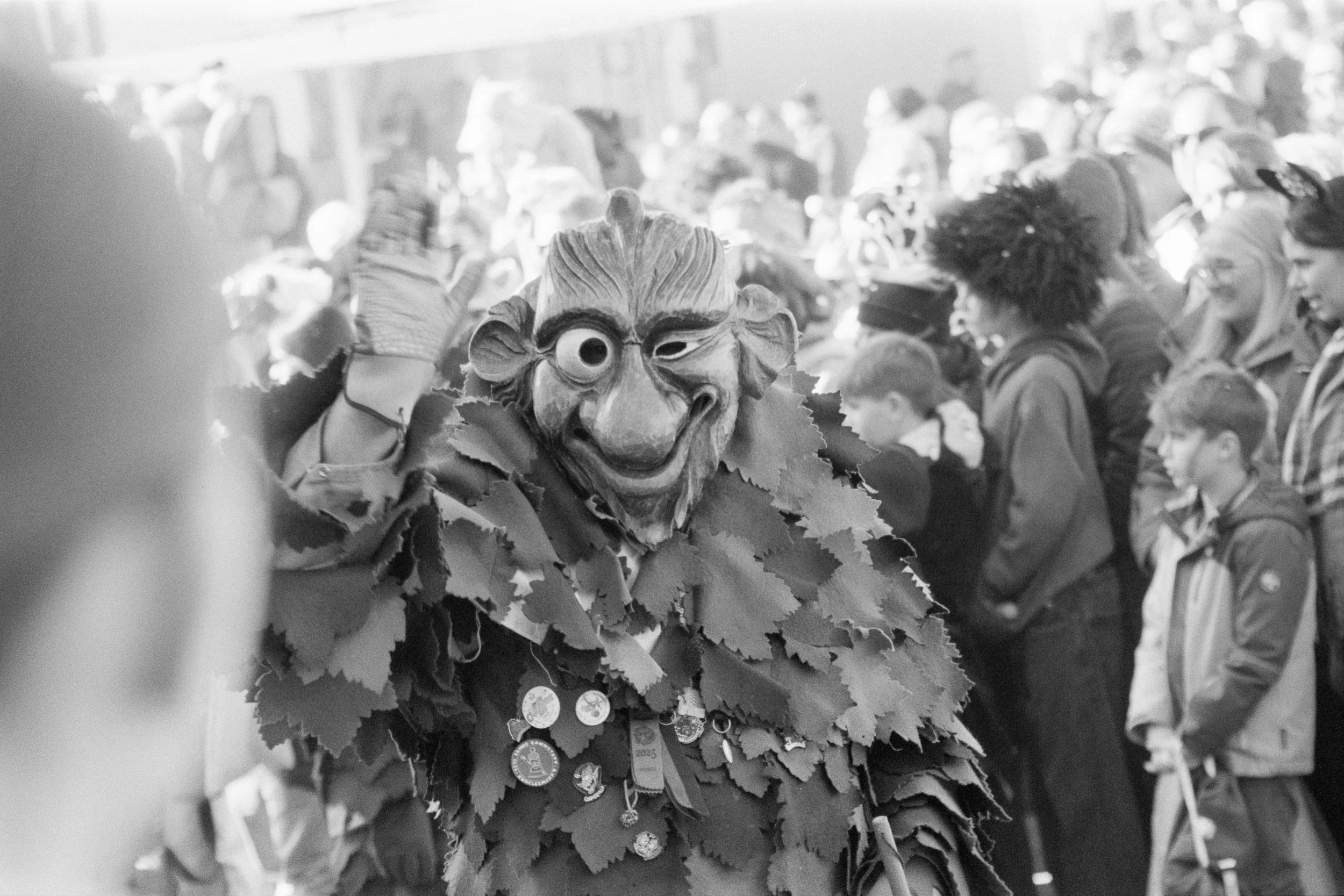 A person dressed in a costume with a large witch or goblin mask and wearing leaves, surrounded by a crowd of people, many of whom are children, in a street parade or festival.