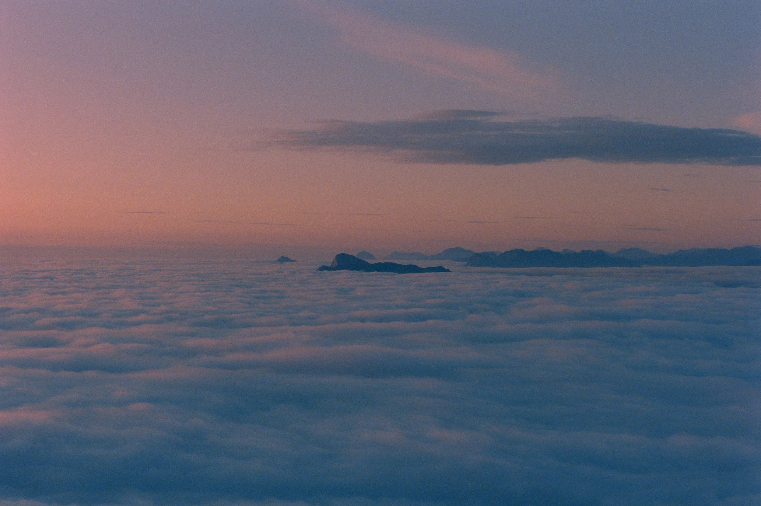 Aerial view of mountains above a sea of clouds at sunset, with the sky displaying pink and purple hues.