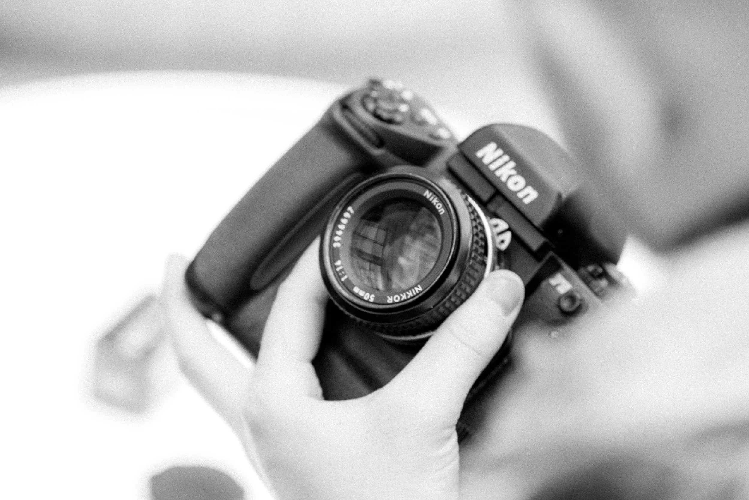 A person holding a Nikon film camera with a close-up view of the camera lens and body in black and white