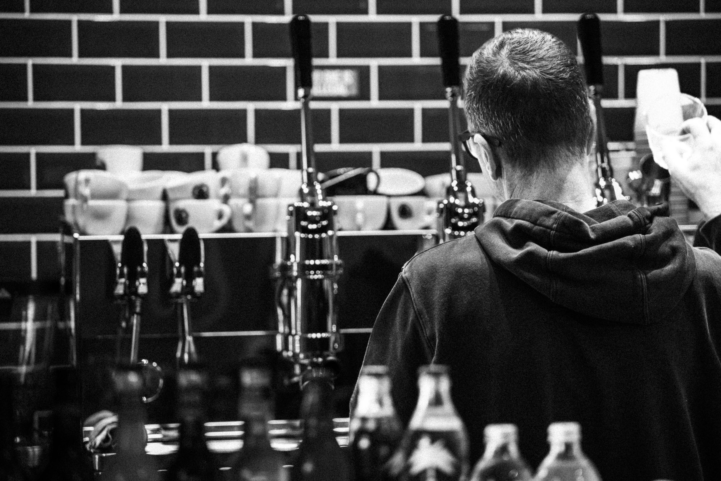 A person with short hair and glasses is standing in front of a wall of coffee cups and bottles, facing away from the camera.