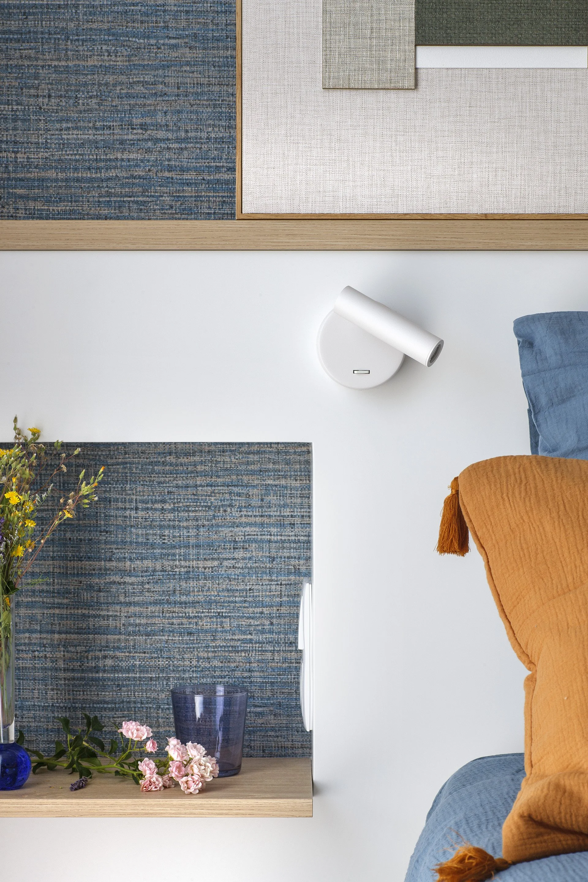 Vista de una sala con pared decorada con paneles de tela en tonos azules y beige, lámpara de pared blanca, mesa con flores y vasos, y parte de un sofá con cojines en colores beige, azul y naranja.