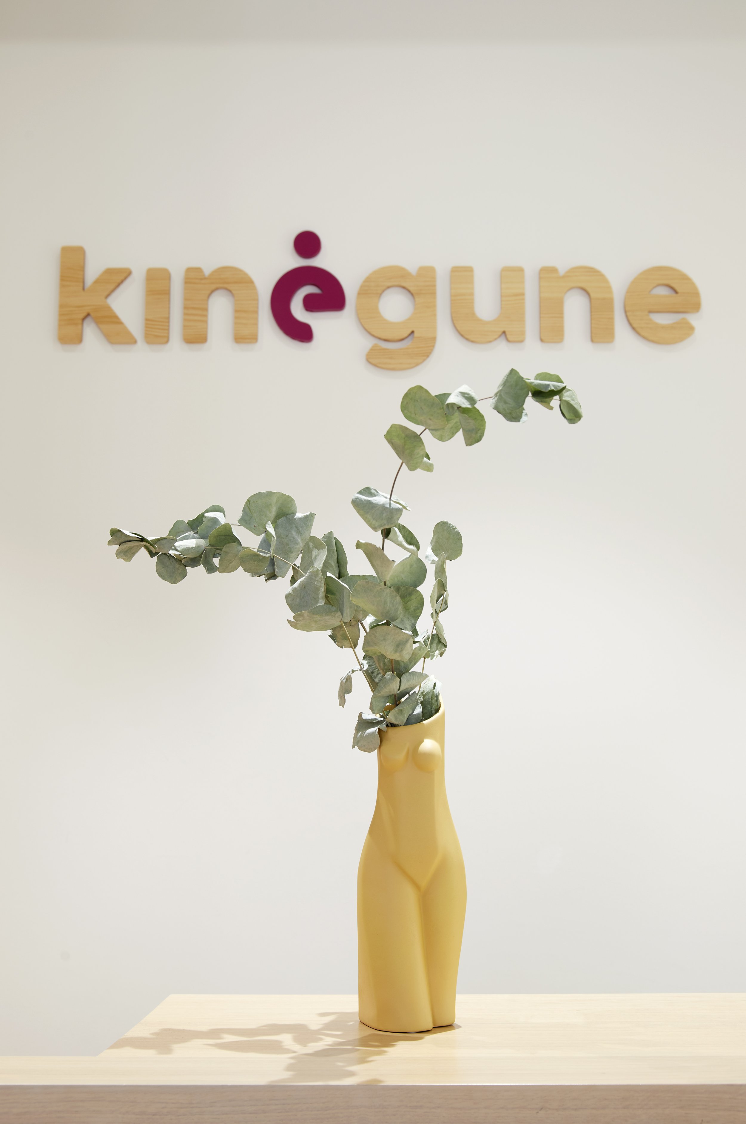 Vaso decorativo en forma de cuerpo humano con plantas en su interior, sobre una mesa de madera clara, con fondo blanco y un letrero en la pared que dice 'kinegune'