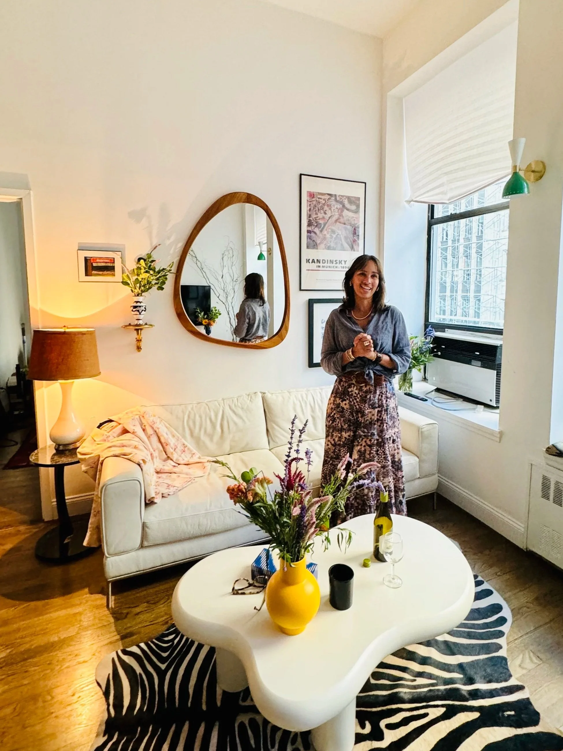 A woman standing in a well-lit living room, smiling, with a white sofa, abstract zebra-patterned rug, and a white coffee table with a yellow vase of flowers, wine bottle, wine glass, and small objects. The room has framed artwork, a mirror, a window with open blinds, and warm lighting.