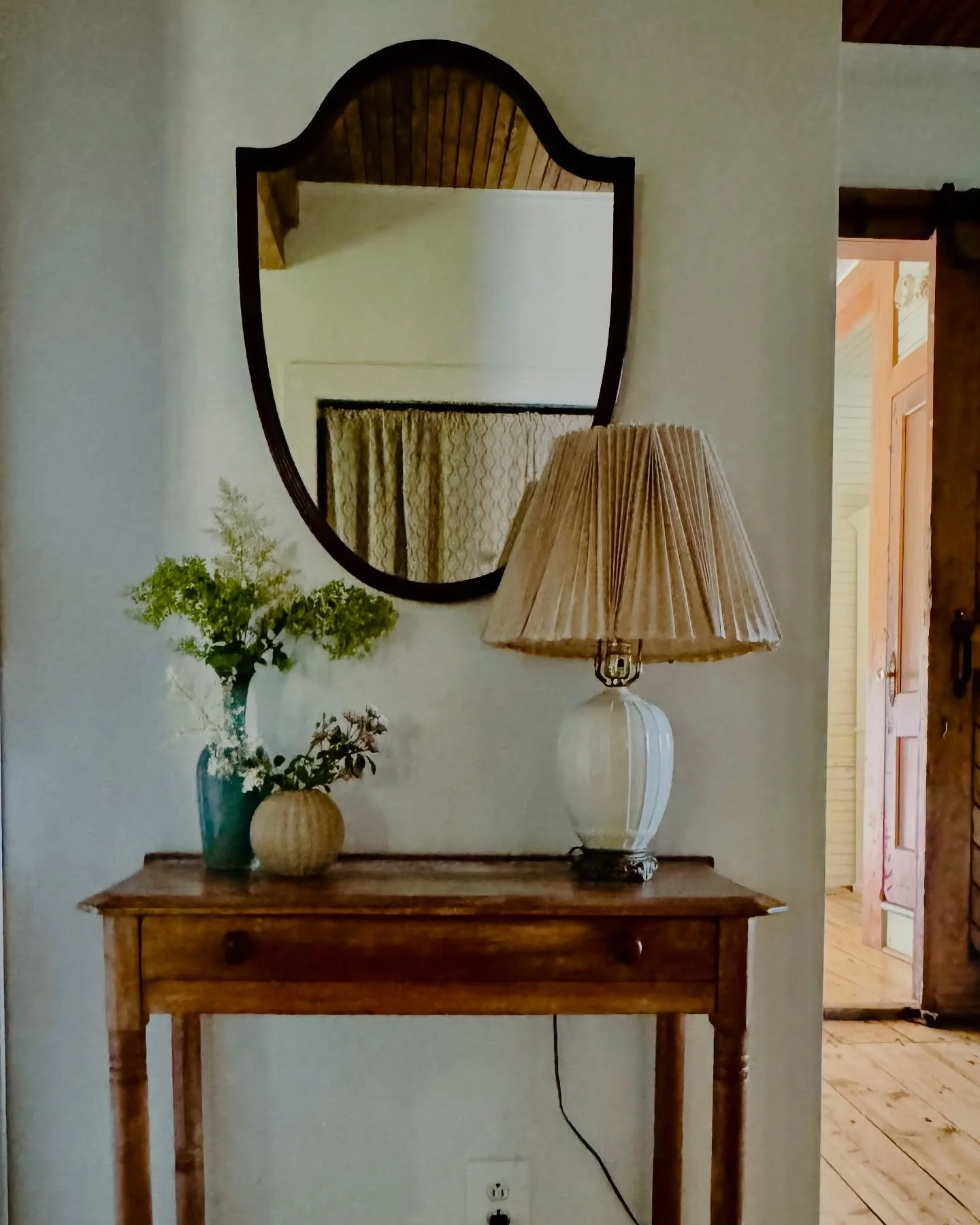 A wooden console table with a lamp, flower arrangement, and mirror on wall behind.