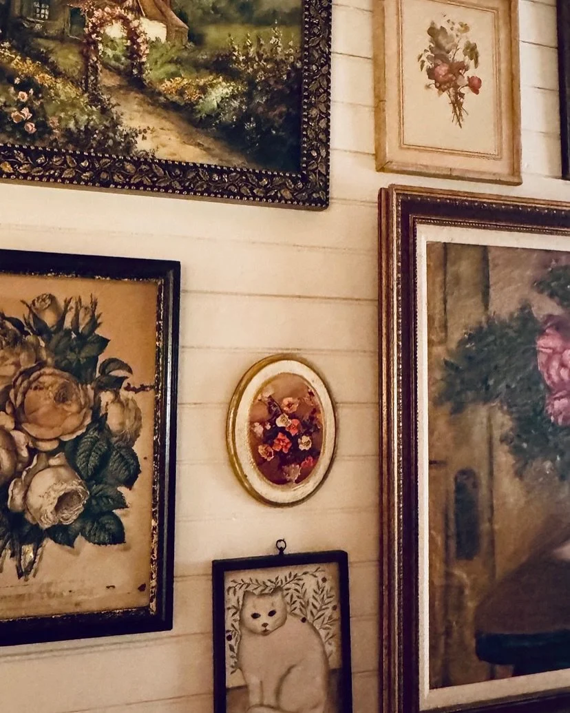 A collection of framed wall art including paintings of flowers, a landscape, an animal portrait, and a floral arrangement, all on a white wooden wall.