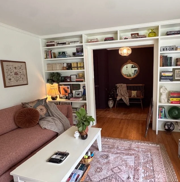 Living room with built-in white bookshelves filled with books and decor, a pinkish sofa with pillows and a blanket, a white coffee table with a potted plant and small decorative items, a rug, and a view into a darker adjacent room with a wooden bench, round mirror, and additional decor.