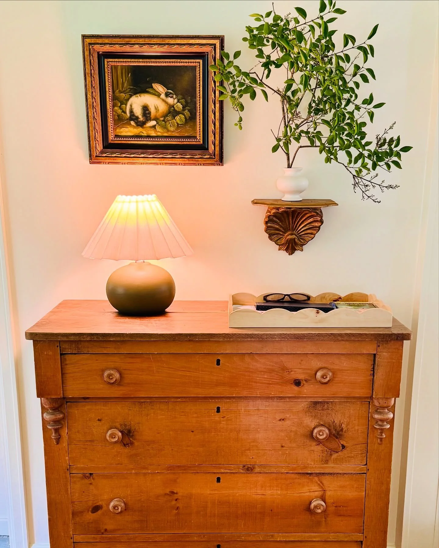 A wooden dresser with a lamp, a tray with glasses and books, a framed rabbit painting, and a plant in a white vase on a wall shelf.