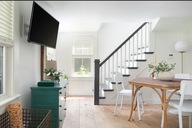 Bright living space with a staircase, a teal dresser, a wall-mounted TV, a window, a round dining table with white chairs, a wicker basket, and plants.