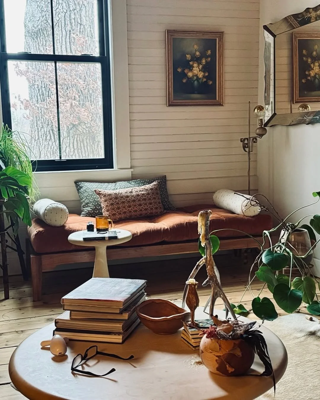 Cozy living room with a wooden bench against the wall, pillows, and a window with a tree outside. A coffee table has stacked magazines, a pair of glasses, and decorative items, including a vase with feathers and natural elements. Wall art decorates the space.