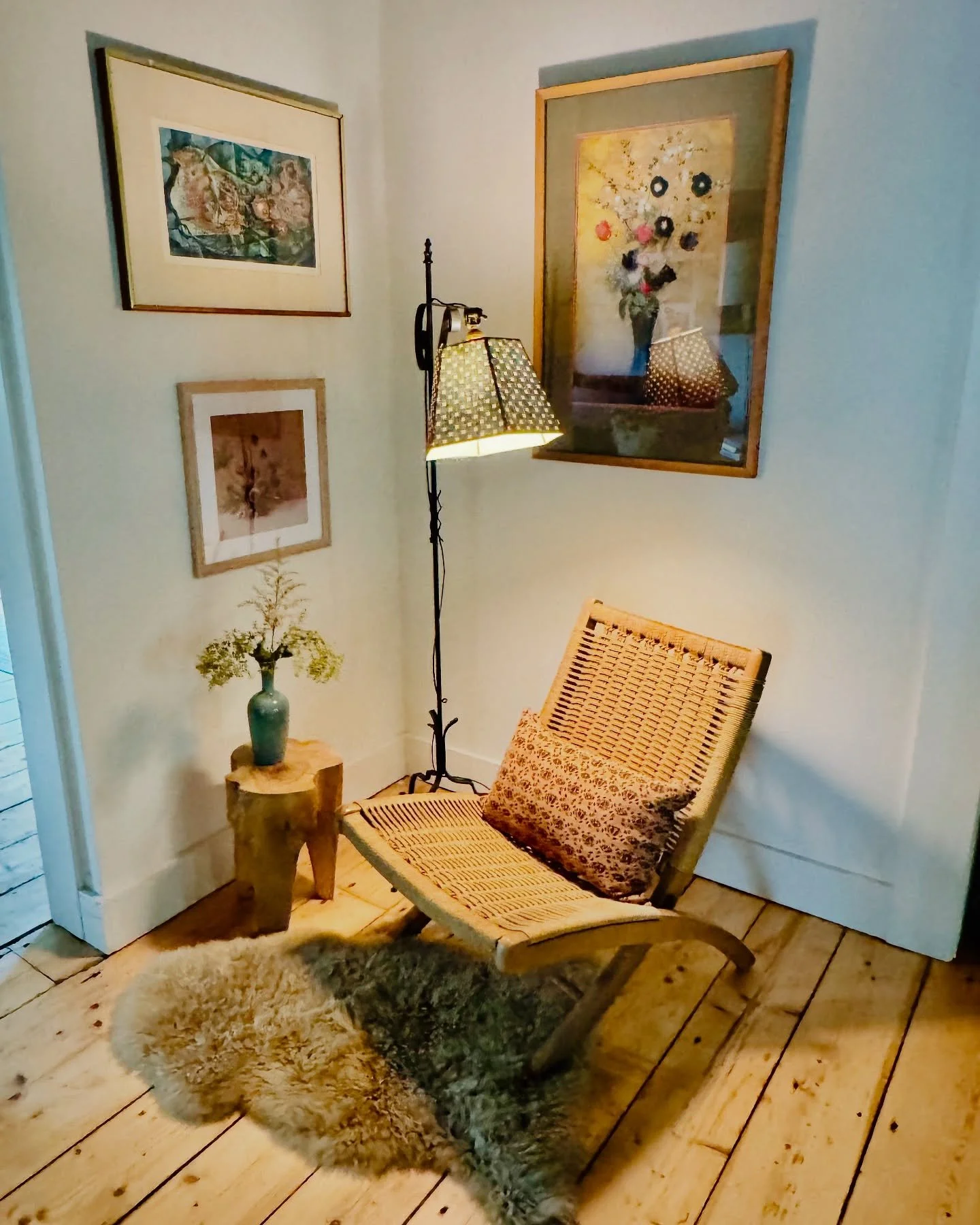 A cozy corner with a woven lounge chair with a patterned pillow, a small wooden side table with a vase of flowers, a standing lamp with a patterned lampshade, and framed artwork on white walls, all on a wooden floor with a faux fur rug.