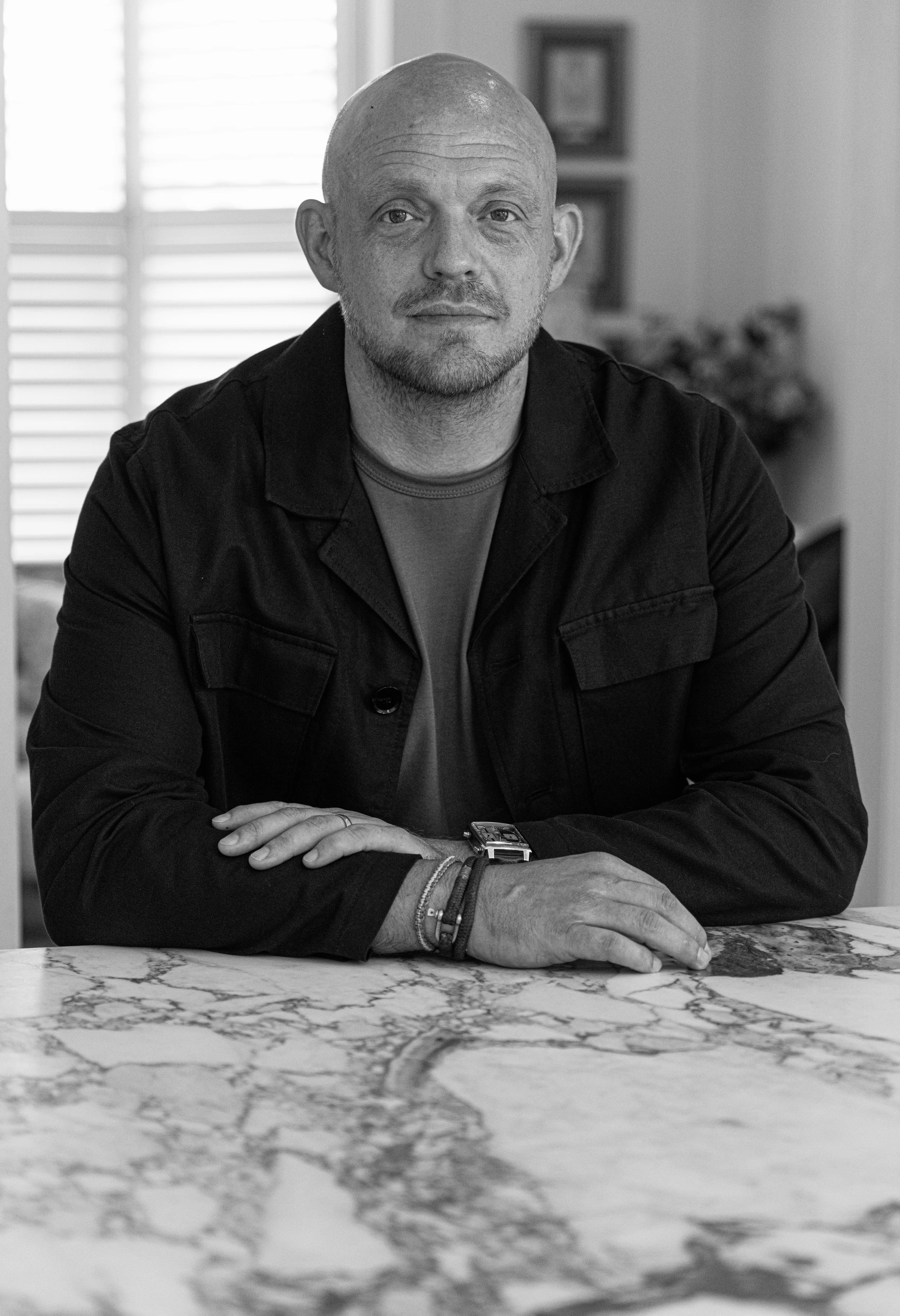 Black and white photo of a man sitting at a marble table, with a window with blinds behind him and picture frames in the background.