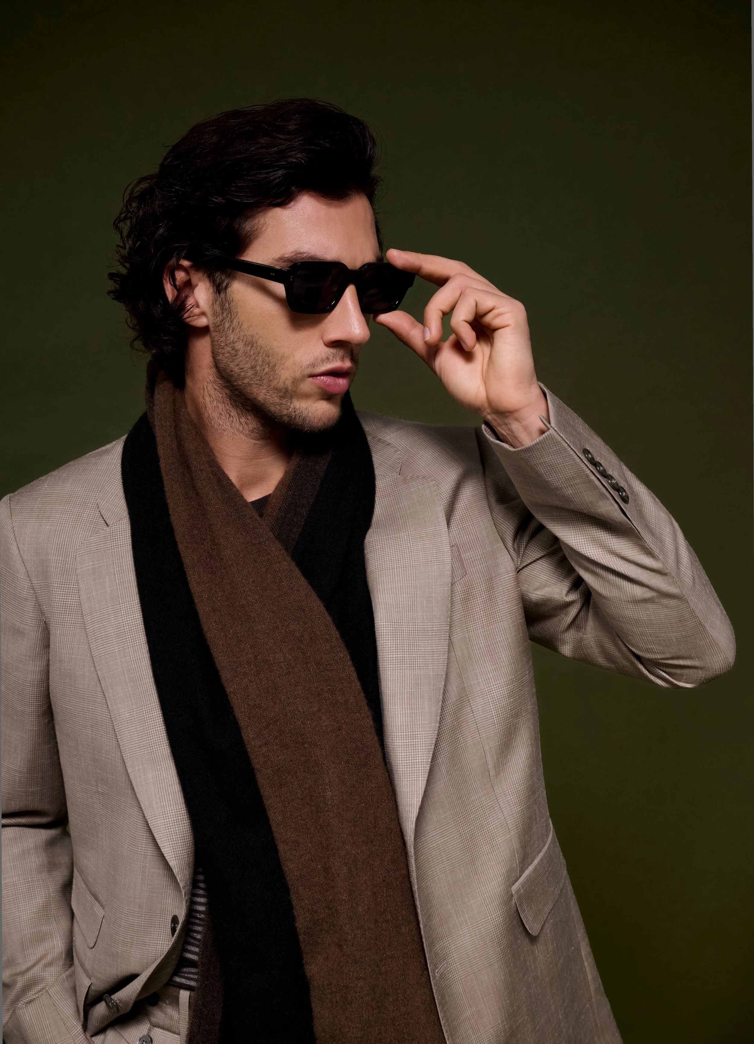 A man with dark, wavy hair, wearing black sunglasses, a beige blazer, a black shirt, and a brown and black scarf, adjusting his sunglasses against a dark green background.