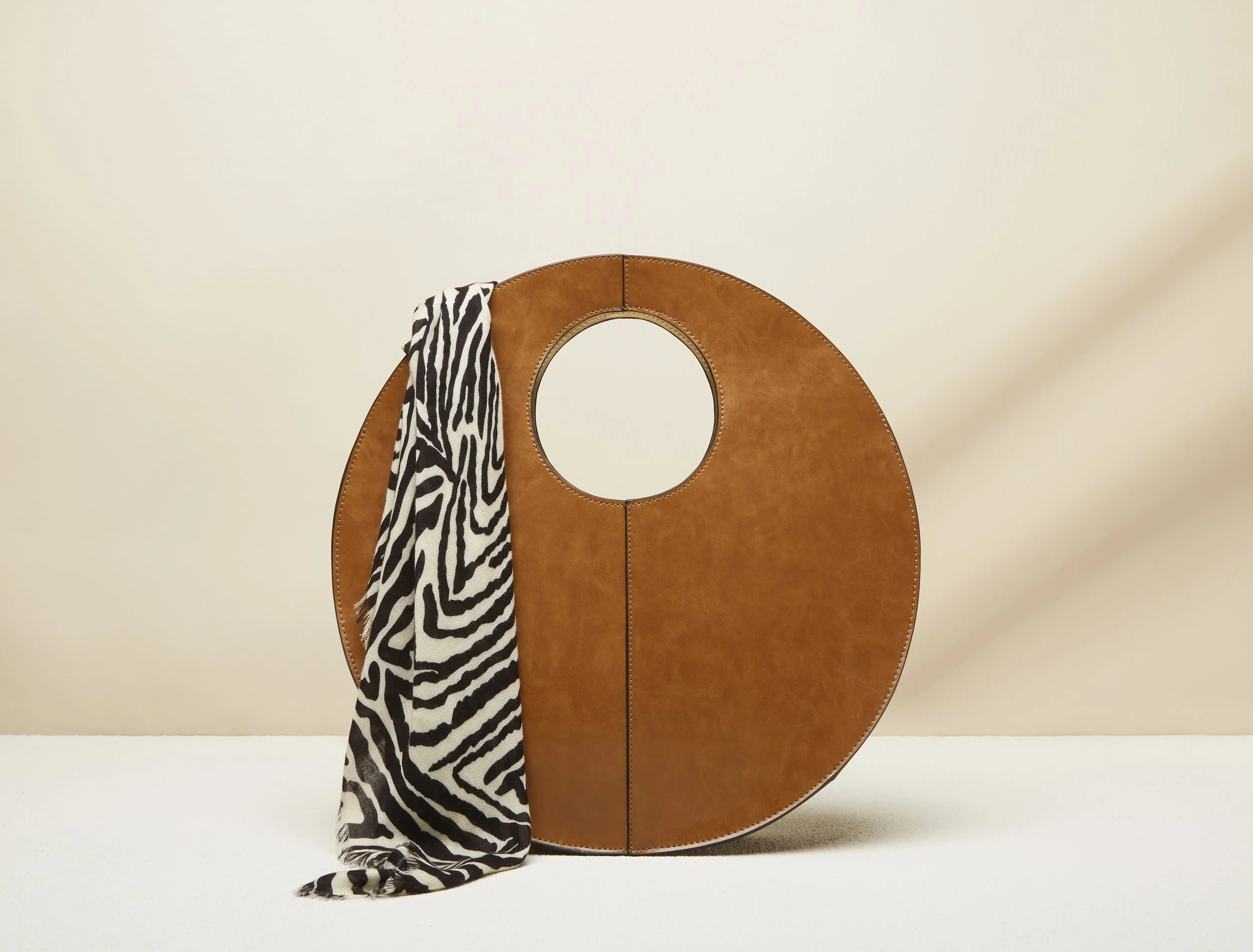 A large, round, leather handbag with a circular handle cutout in the center, placed on a surface against a plain background, with a zebra-print scarf draped over its left side.