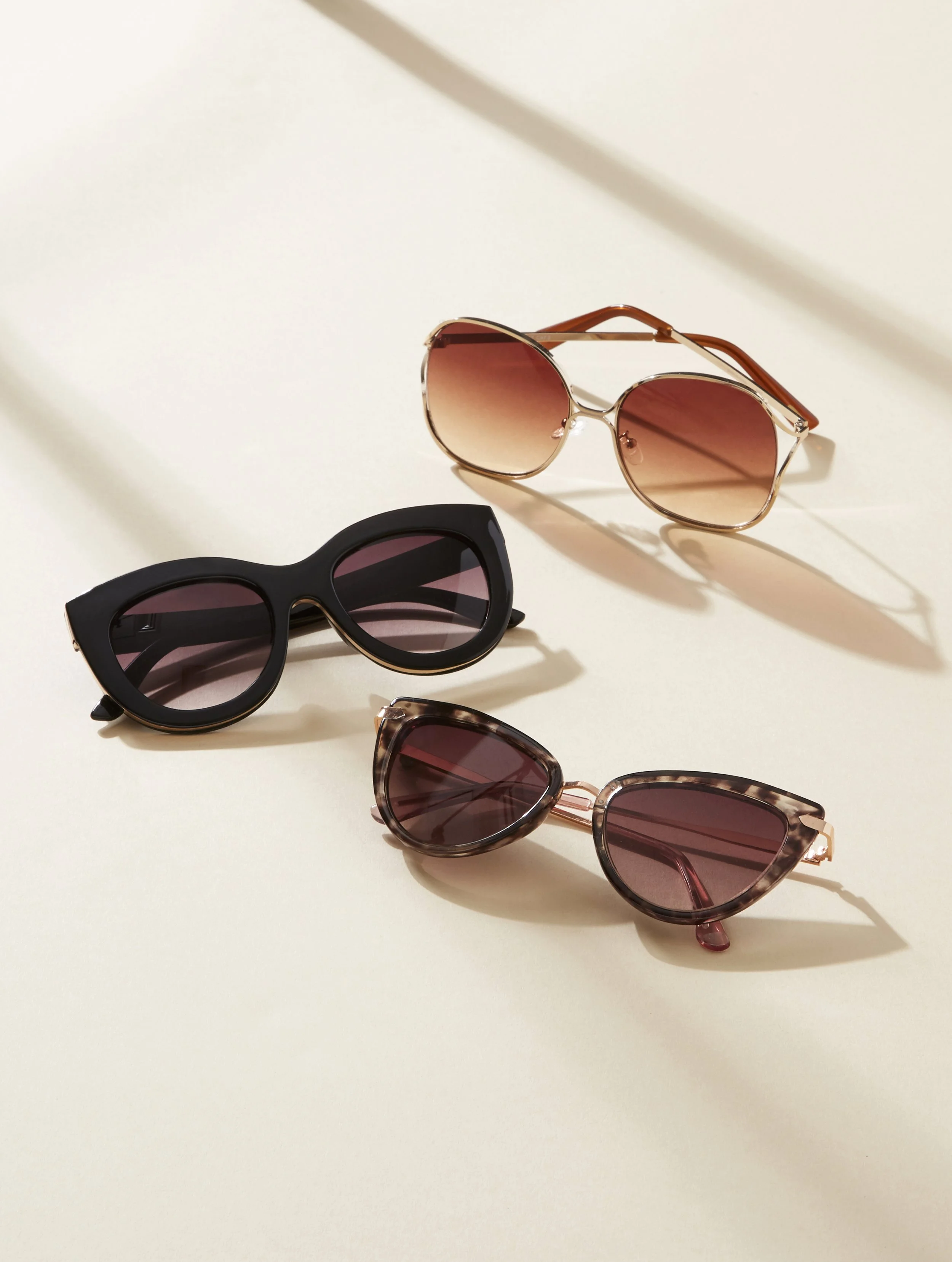 Three pairs of sunglasses with different styles and colors on a plain white surface, with shadows cast by the sunlight.