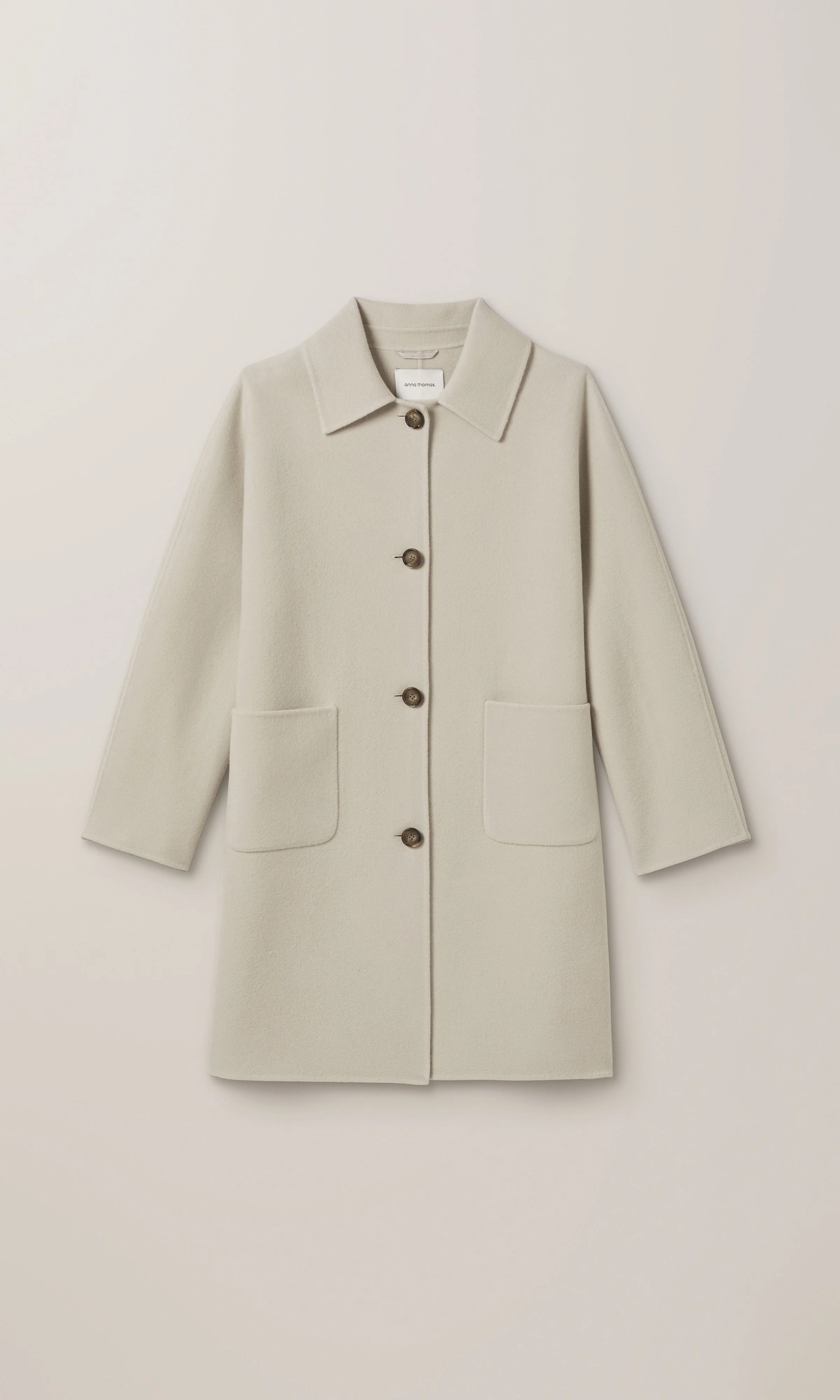 A cream-colored coat with a collar, buttons down the front, and two front pockets, hanging on a white wall.