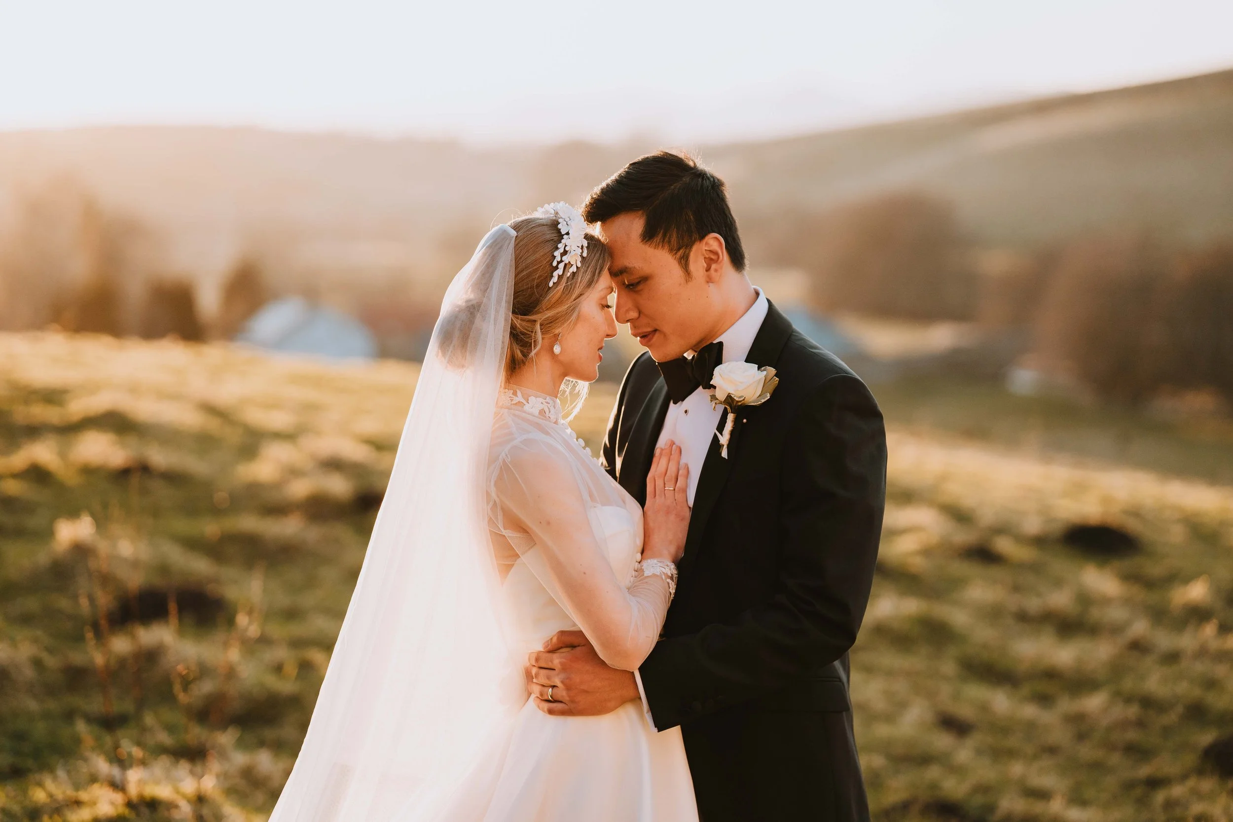 A newlywed couple embracing outdoors during sunset, with eyes closed and foreheads touching, in a peaceful, rural landscape.
