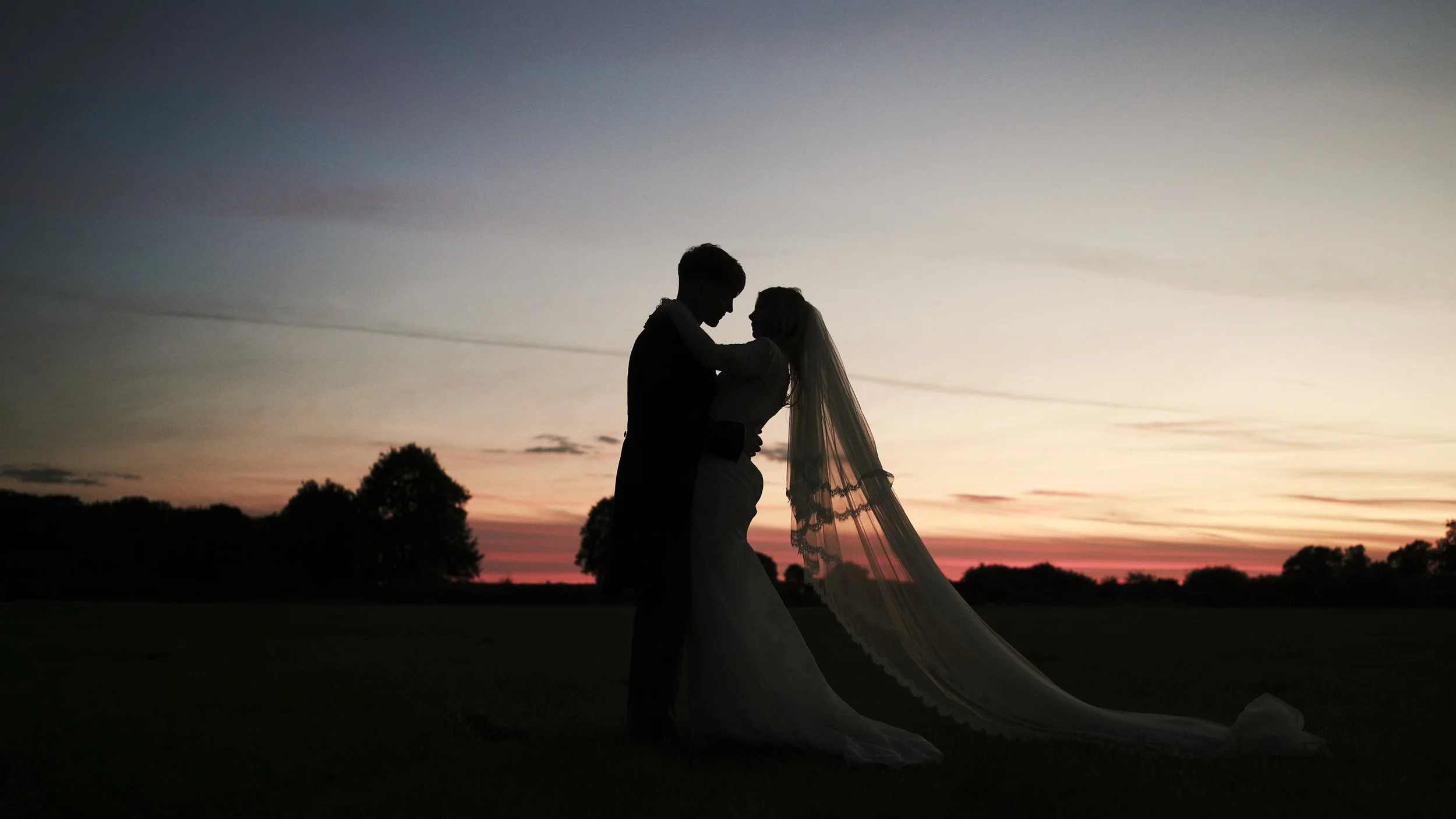 Silhouette of a bride and groom standing close together during sunset, outdoors with trees in the background.