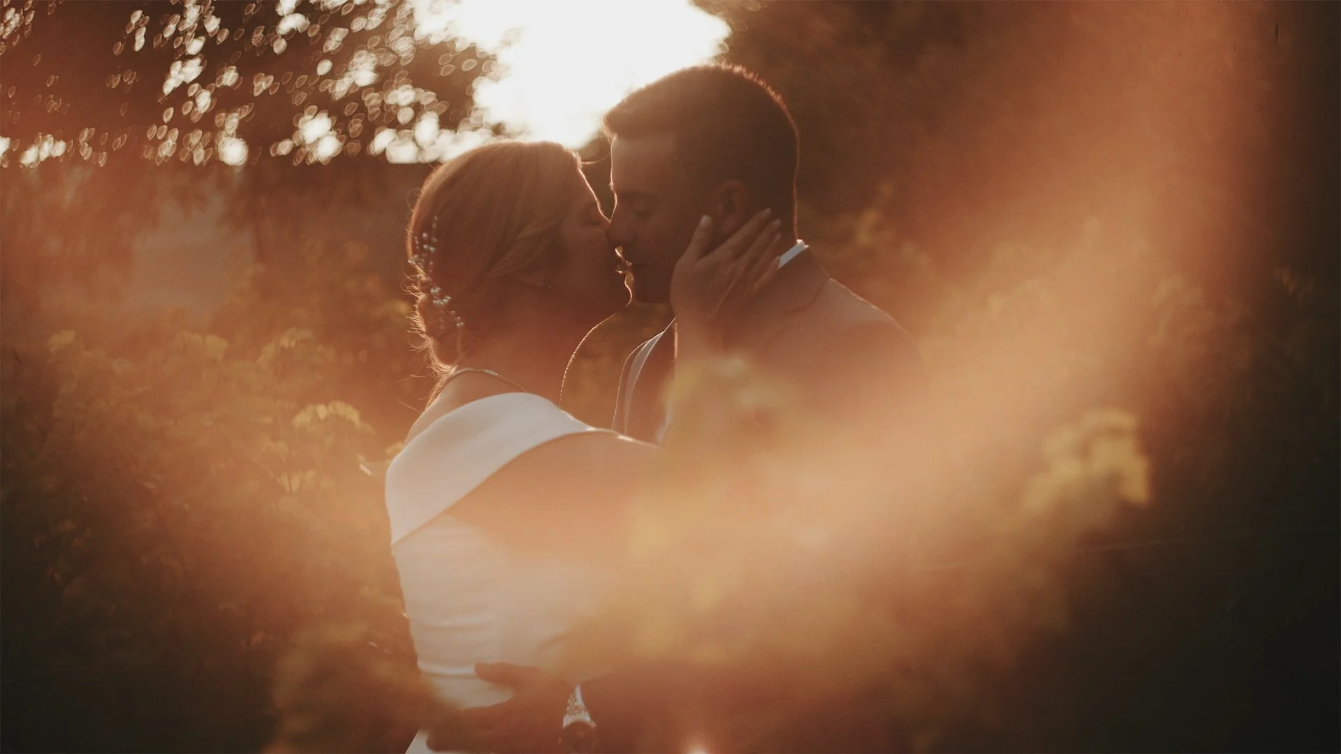 A couple kissing outdoors during sunset, surrounded by warm lighting and soft focus foliage.