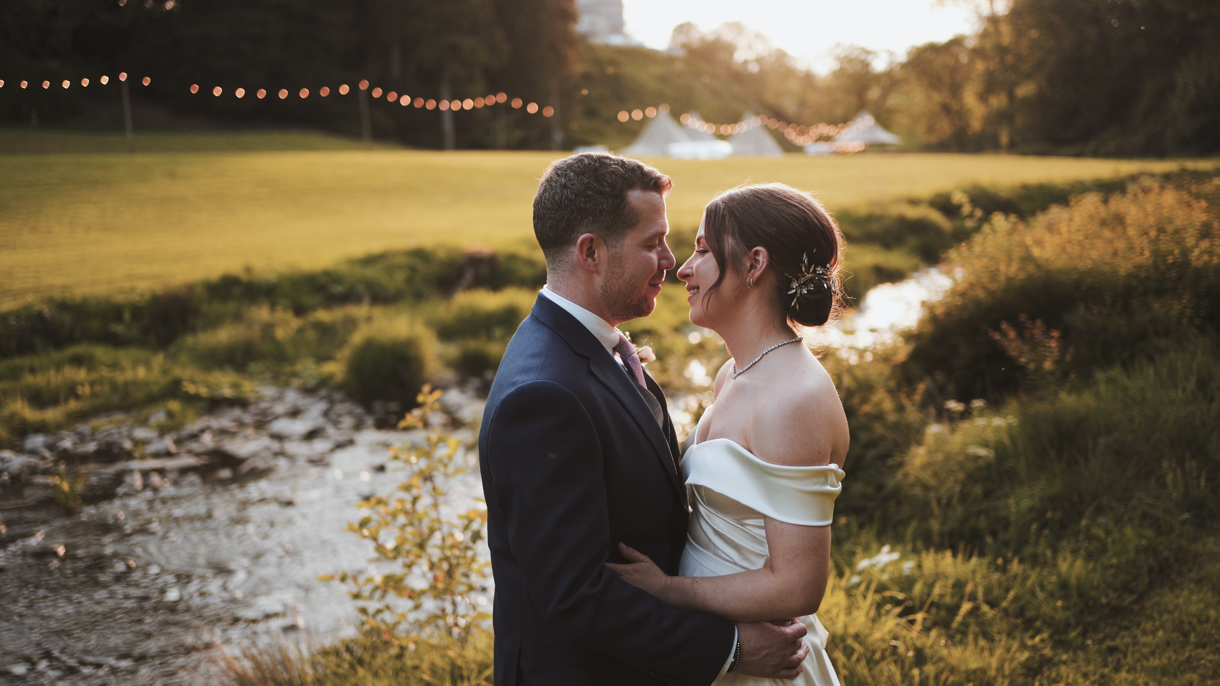 A bride and groom standing close together outdoors at sunset, with a scenic background of a pond, greenery, and string lights.