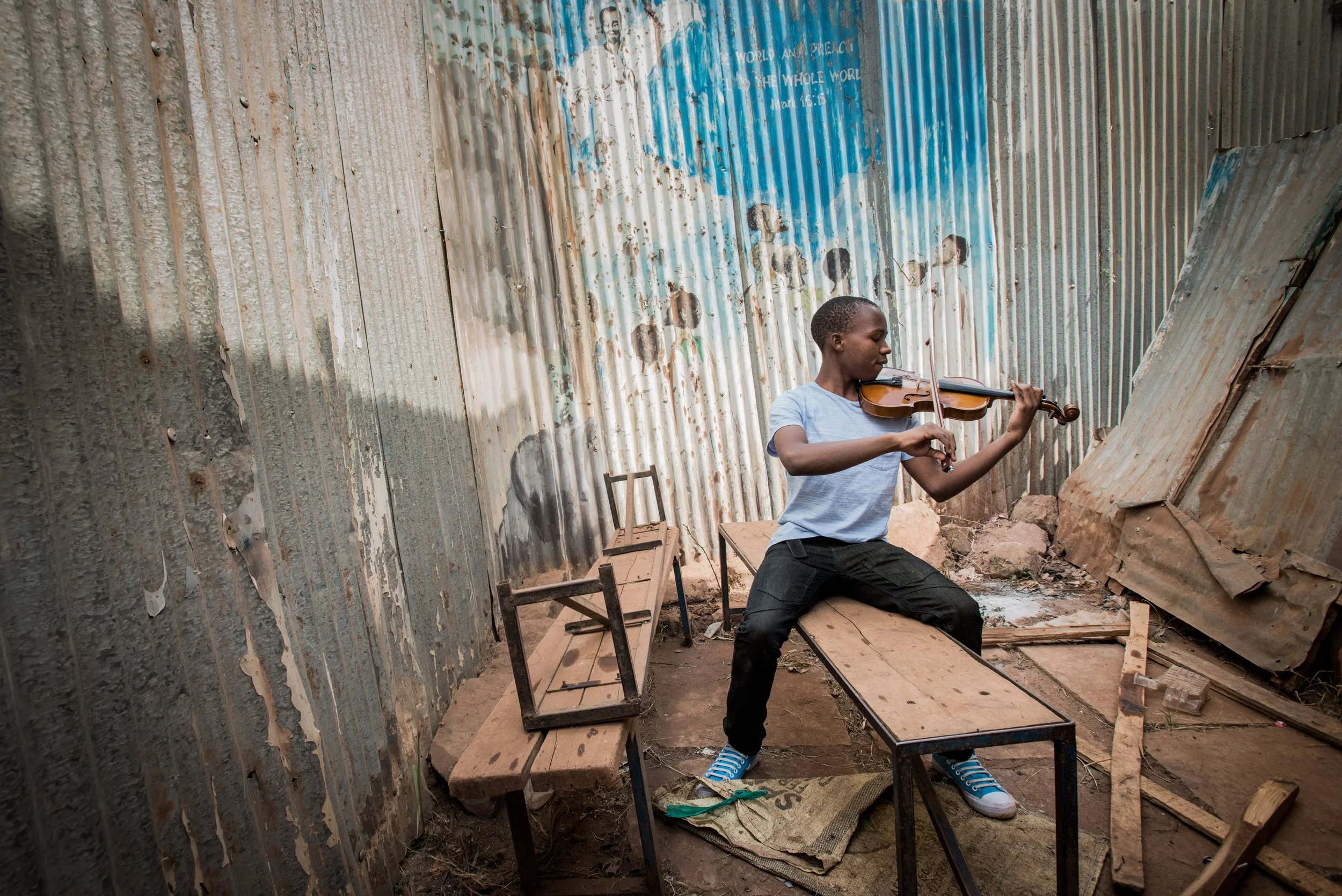 A young boy playing the violin while sitting on a wooden bench inside a makeshift structure with corrugated metal walls, with a mural painted on the wall behind him depicting children.