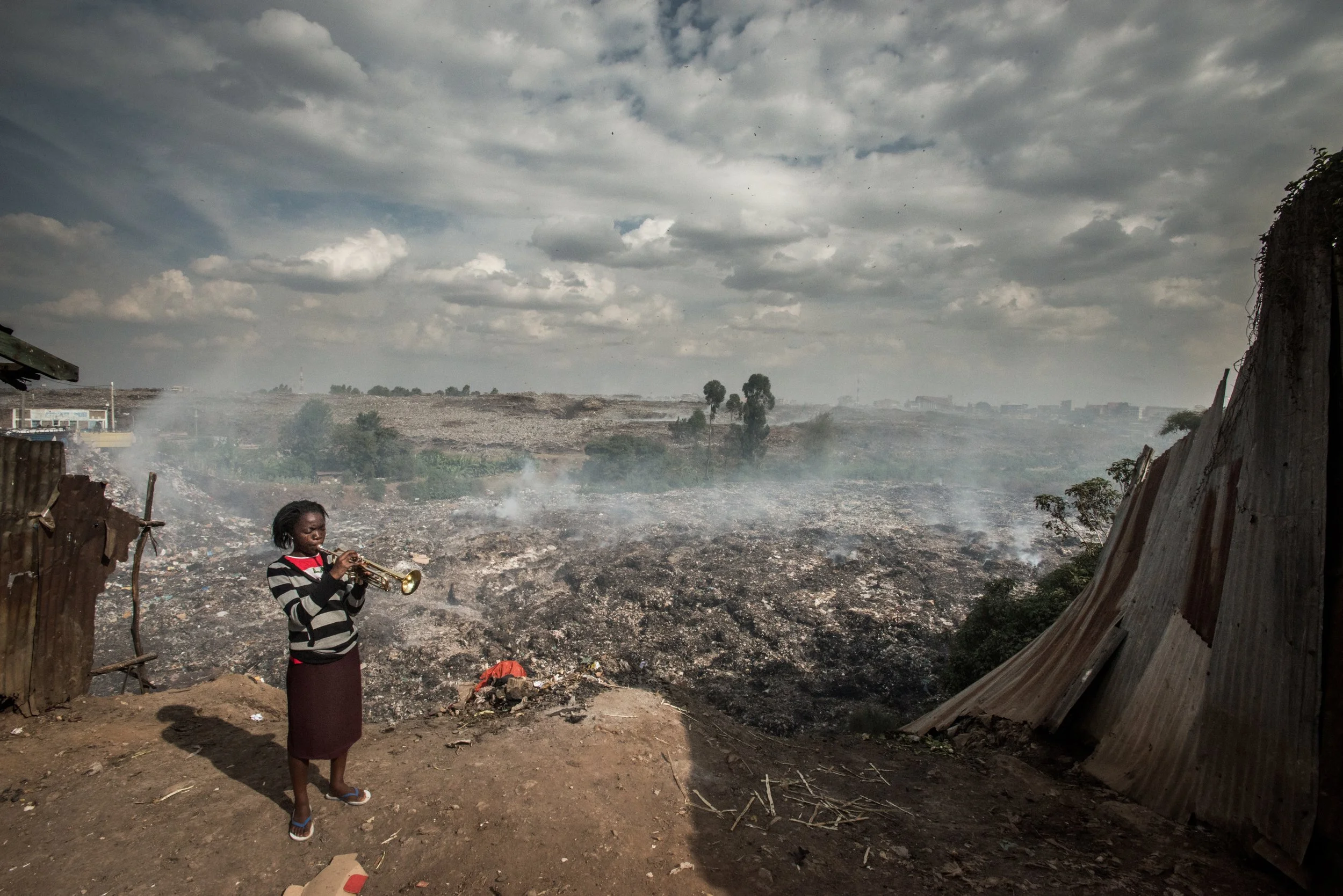A young girl standing on dirt holding a trumpet with a vast dump site and smoke in the background under a cloudy sky.