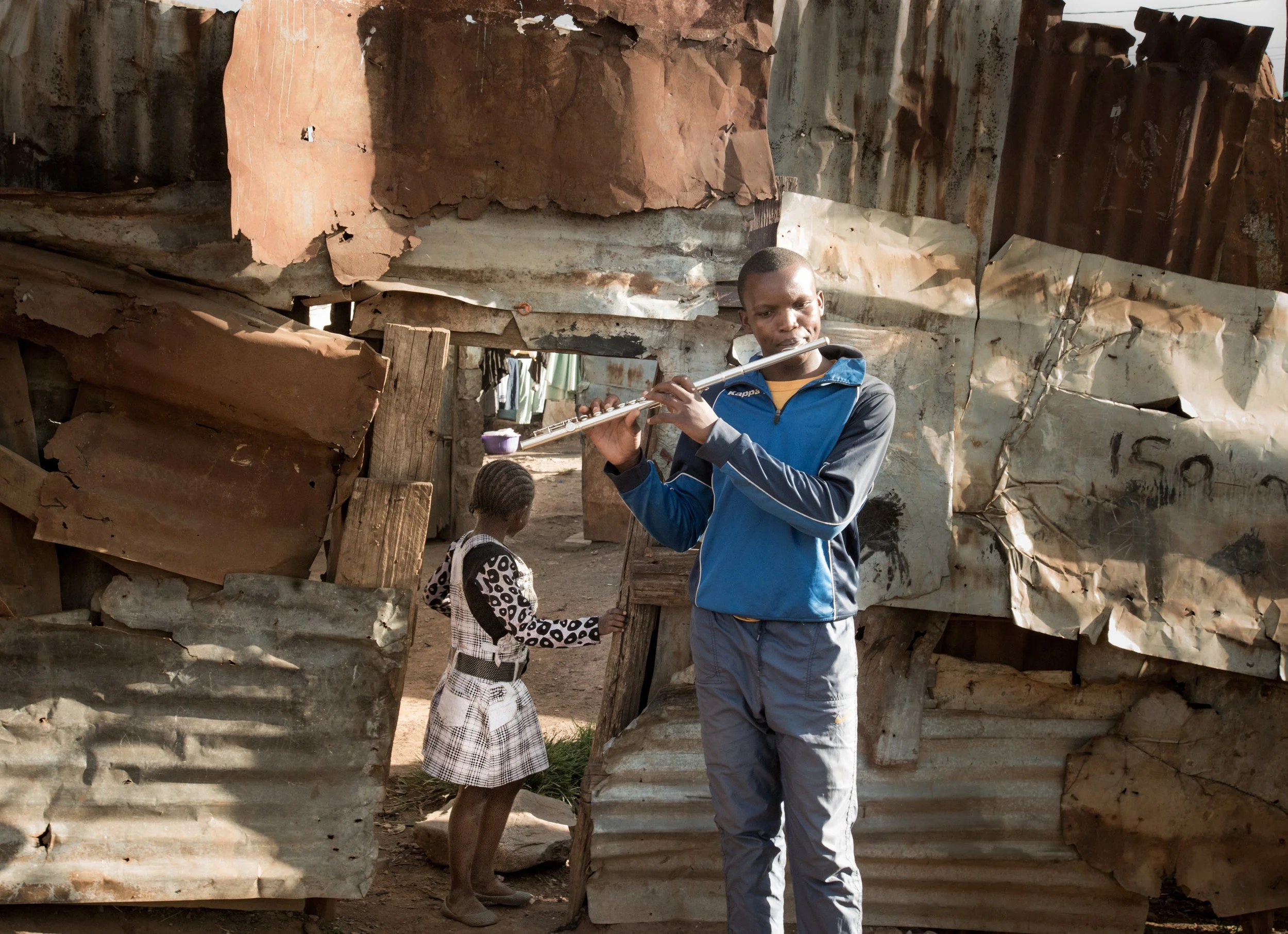 A young man playing a flute in a makeshift shelter made of corrugated metal sheets, with a young girl in a dress standing nearby in a seemingly impoverished area.