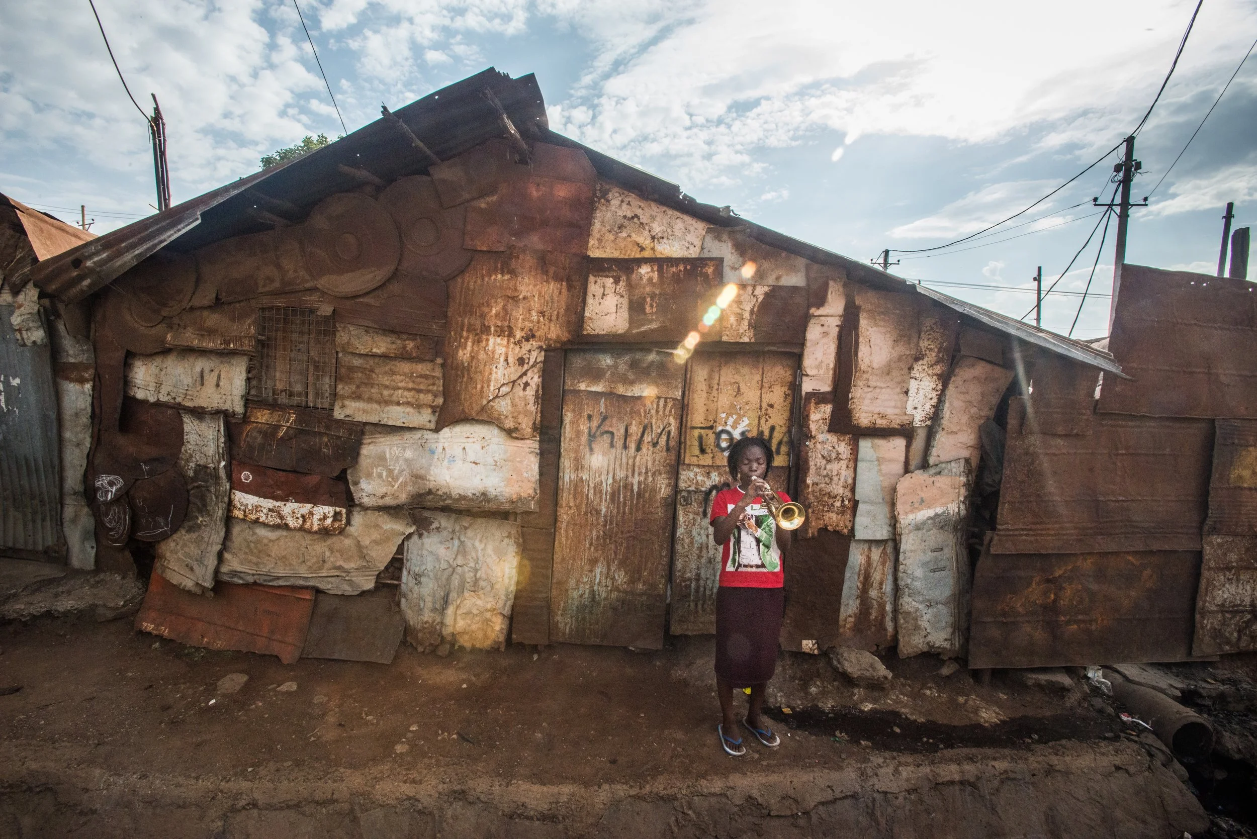 A young girl in a red shirt playing a trumpet in front of a makeshift shack built from various rusted metal sheets.