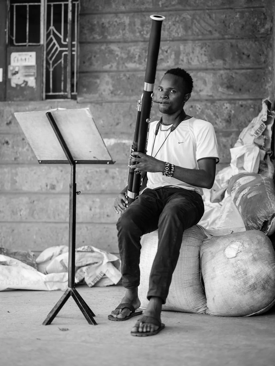 A man sitting on a cushion playing a long wind instrument, with a music stand and a brick wall background.