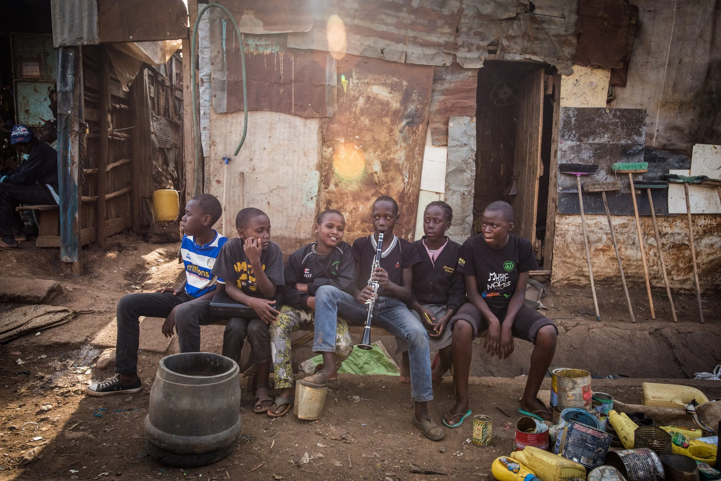 Six children sitting on a bench outside a makeshift building made of wood and metal, with various objects and cans on the ground in front of them, one child playing a clarinet.