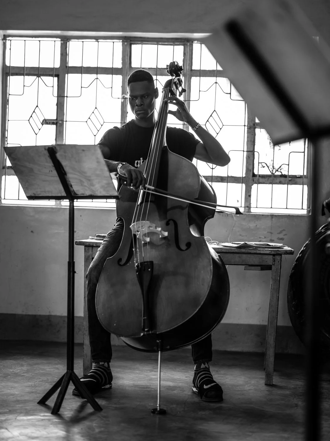 A young man playing a double bass in a room with barred windows, music stand, and a table.