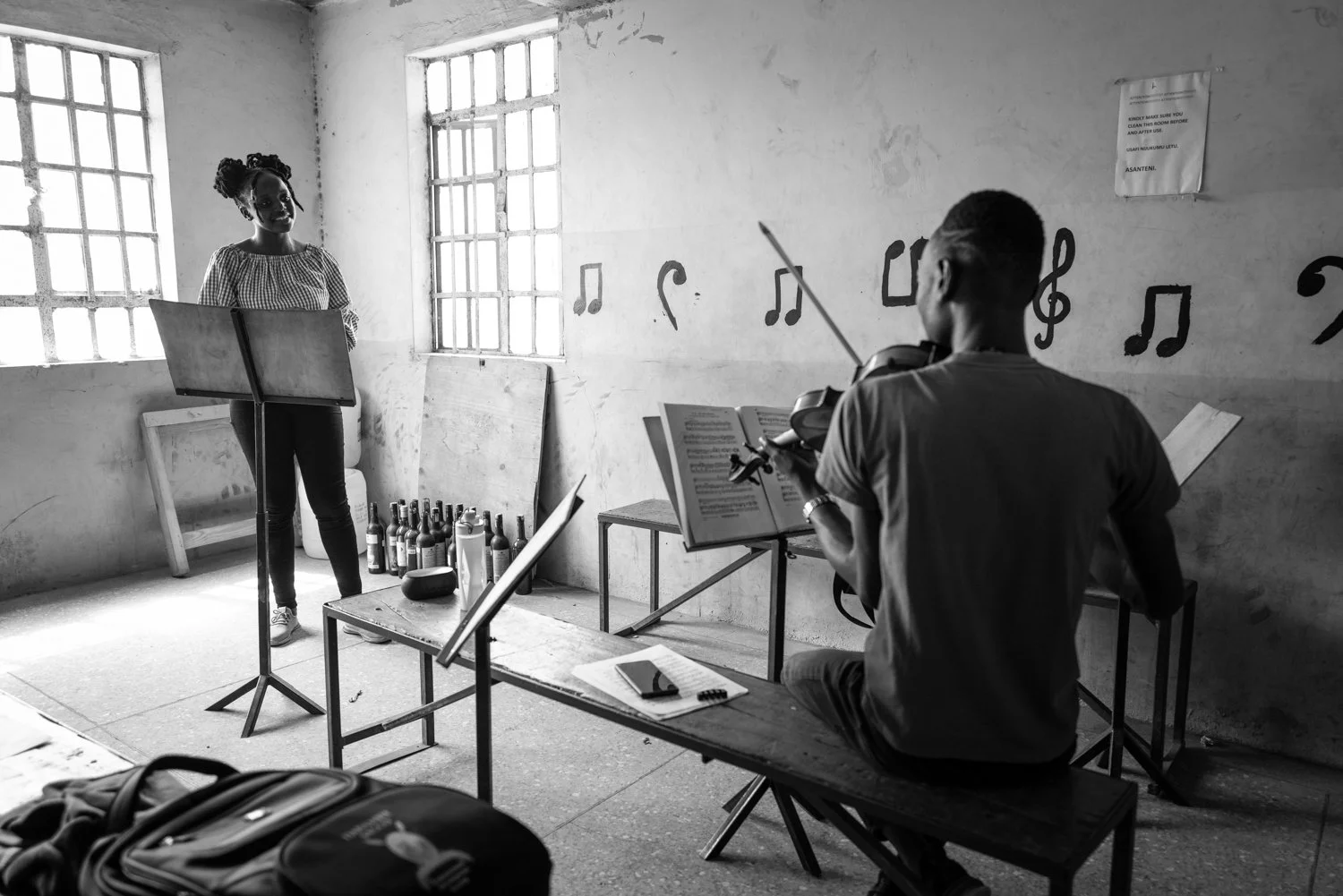 A woman standing in front of a music stand and a man sitting on a bench playing the violin in a room with musical notes painted on the wall.