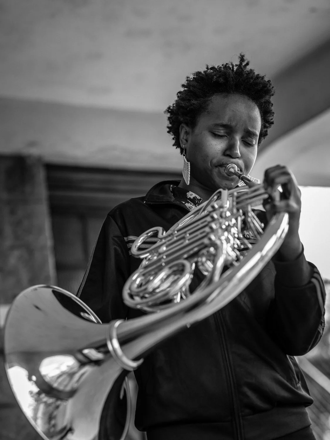 A woman playing a wind instrument, likely a French horn, with her eyes closed, wearing earrings and a dark jacket.