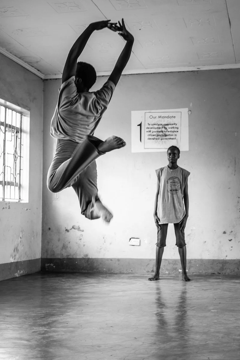 A young person is performing a high jump or dance move indoors, with another person standing nearby watching, in a room with plain walls and a window.
