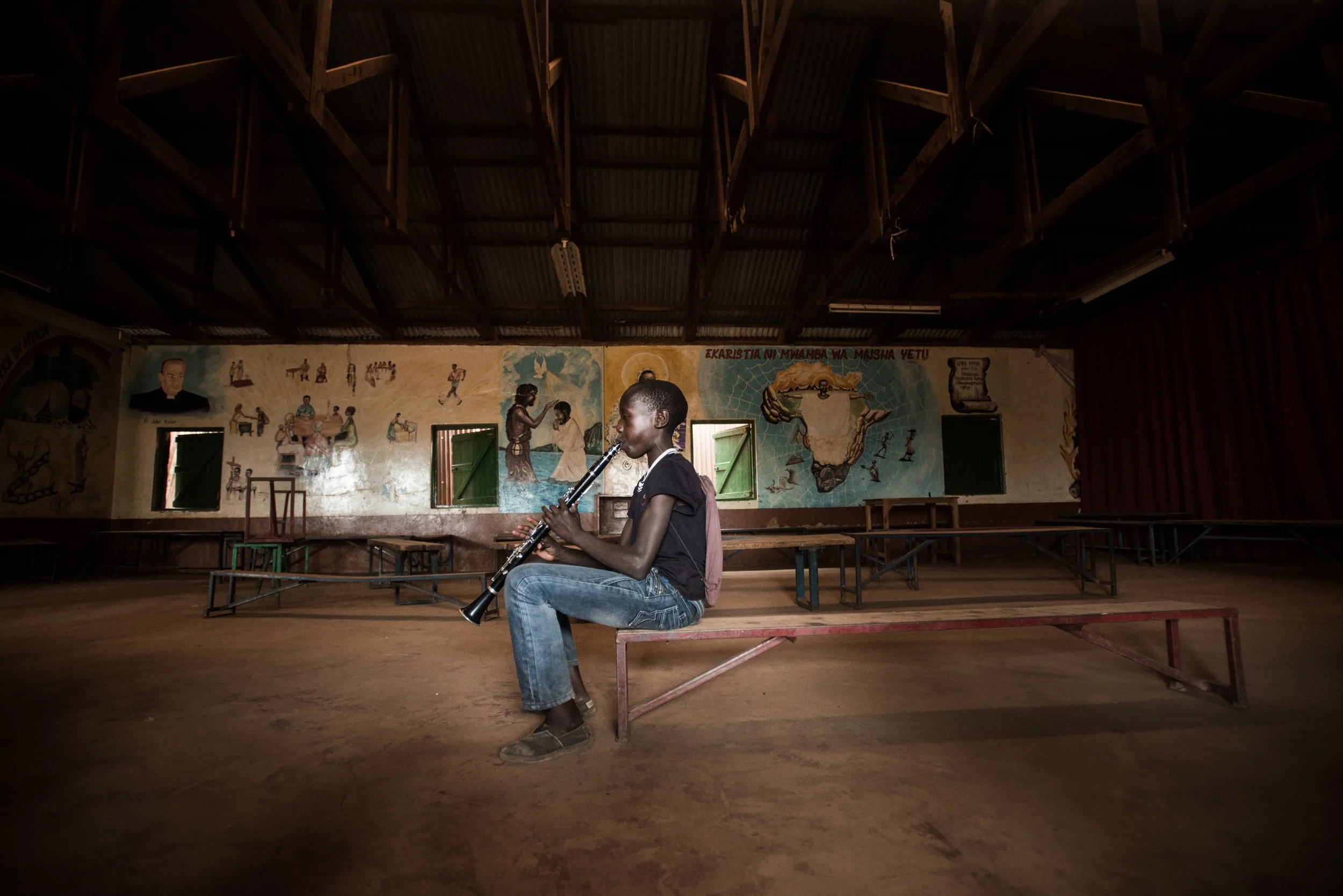 A young person sitting on a bench inside a large, dimly lit room, playing a clarinet. The room has a wooden ceiling and walls decorated with murals and artwork.
