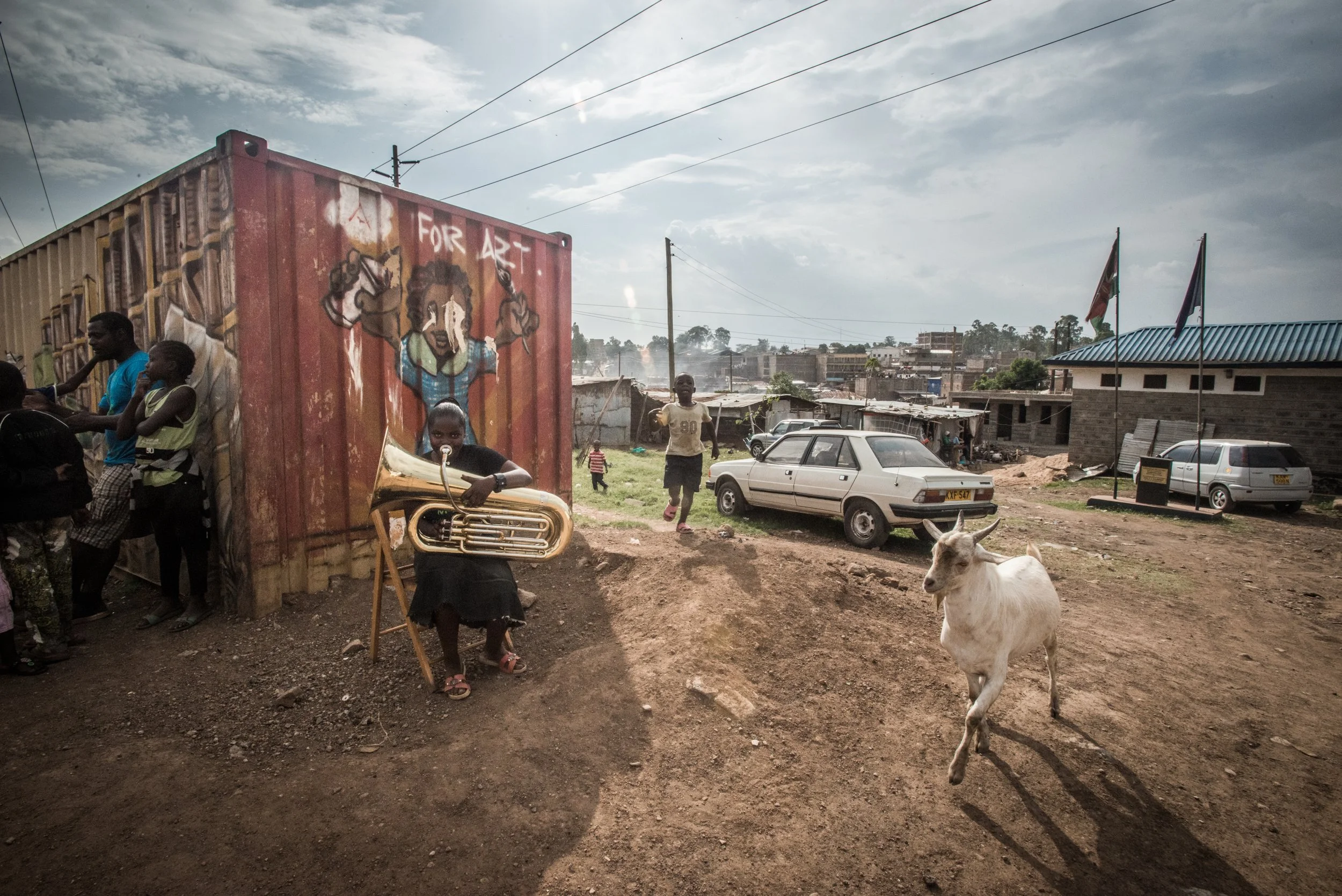 A young girl sitting and playing a tuba in front of a colorful mural with the words "For Art" on a red wall; children and adults gather around nearby; a goat runs across a dirt area; a white car is parked behind the girl; in the background, there are