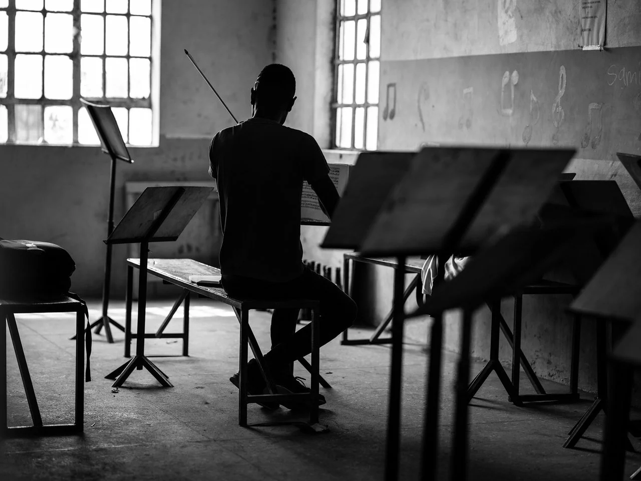 A person sitting in a music classroom with music stands and sheet music, facing large windows and a chalkboard wall.