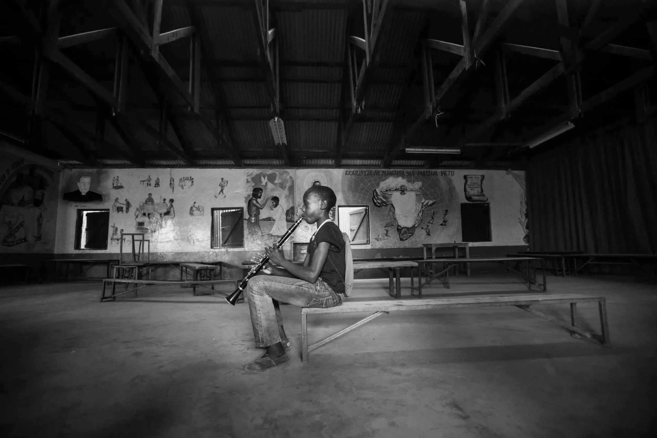 A young boy sitting on a bench inside a spacious room playing a clarinet. The room has a high ceiling supported by wooden beams and large murals painted on the walls, depicting various scenes and figures.