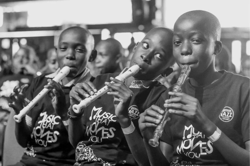 Three children playing flutes at an indoor event, wearing matching T-shirts.