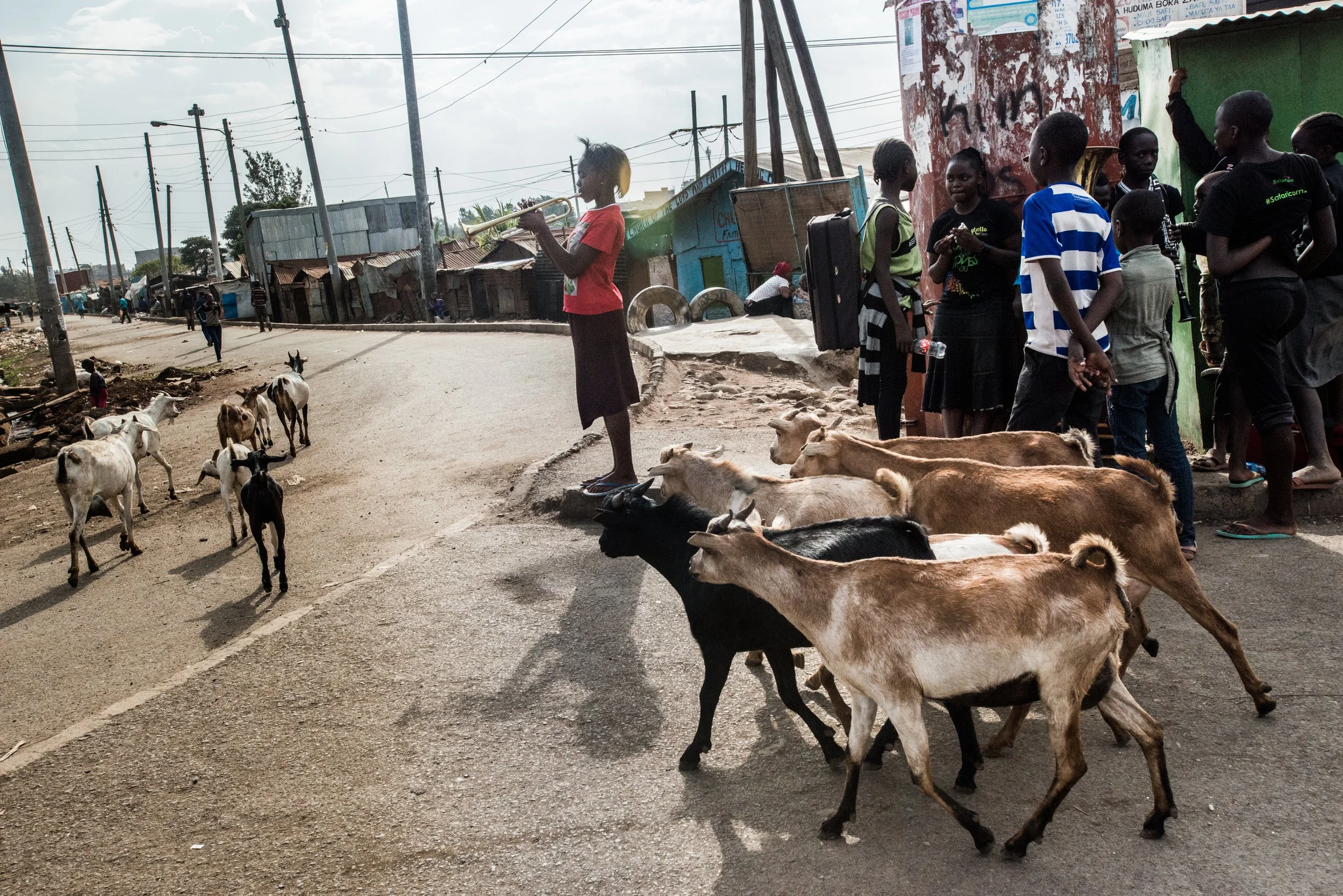 A group of children and goats on a dirt street in a rural area, with some children watching and playing near a green building, and a girl in red standing on a rock playing a trumpet.