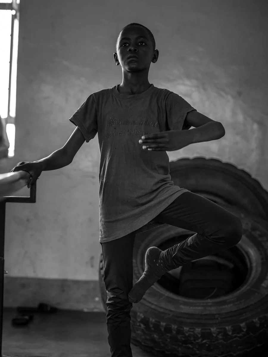 A young boy practicing a dance move or yoga pose indoors, balancing on one leg with one arm extended, in a room with large tires in the background.