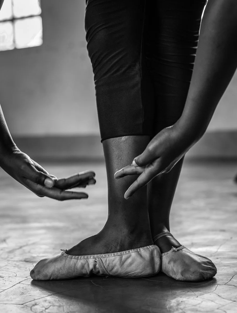 Ballet dancer's feet in pointe shoes, with instructor adjusting the shoes in a dance studio.