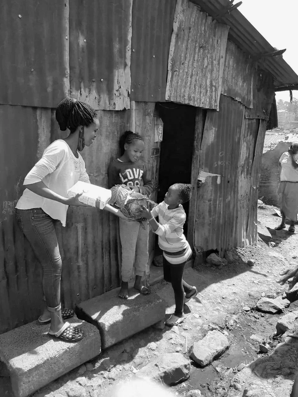 A woman on a small step handing a package to a woman at a doorway while a young girl stands nearby looking on, in front of a makeshift wooden house. A person in the background appears to be taking a photo.
