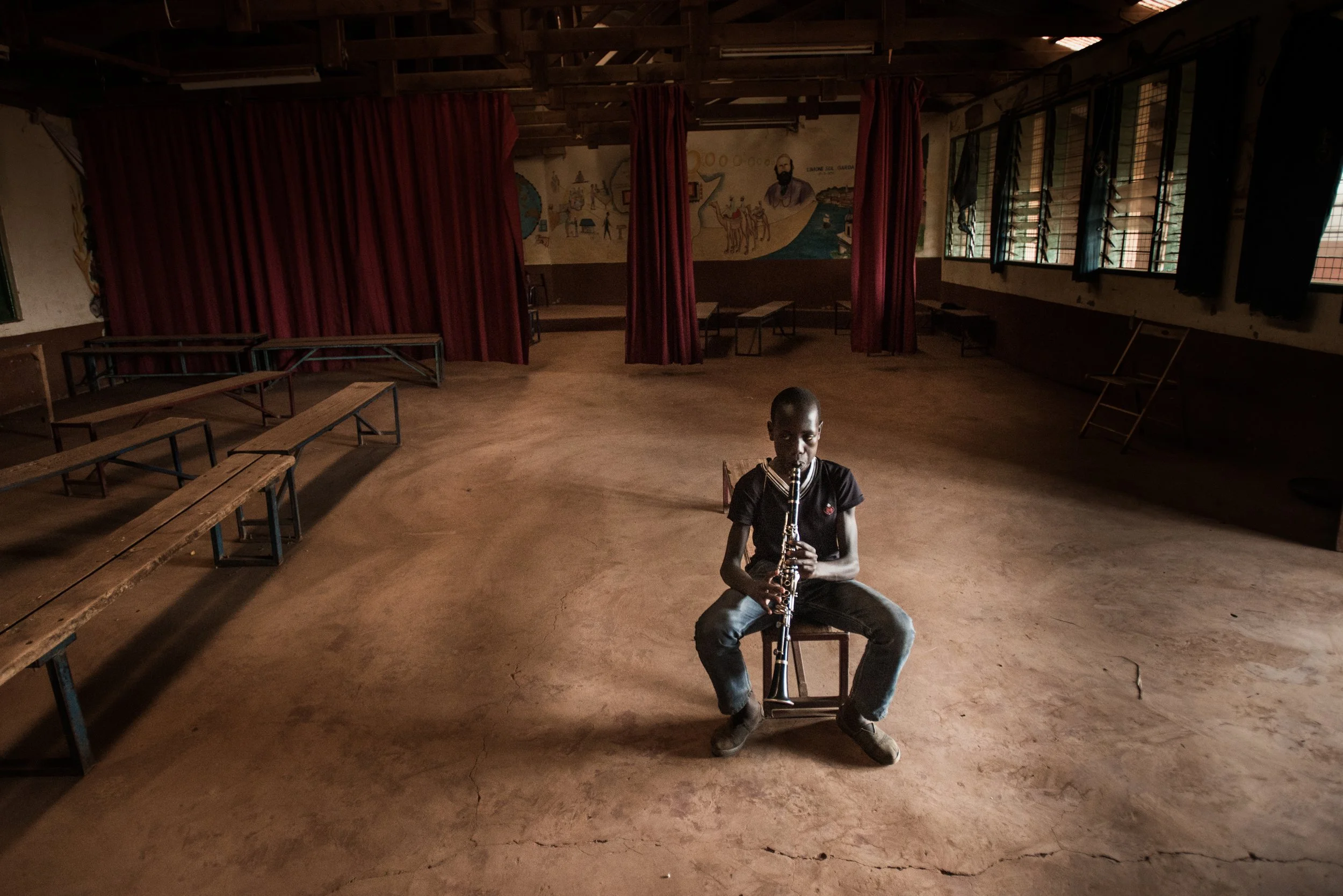 A young boy sitting on a chair in a large, empty indoor room, playing a clarinet. The room has red curtains, wooden benches, and windows with open louvers, with a mural on the back wall.