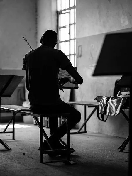 A person sitting on a bench in an art studio, with a paintbrush attached to their back, working at a table near a large window.
