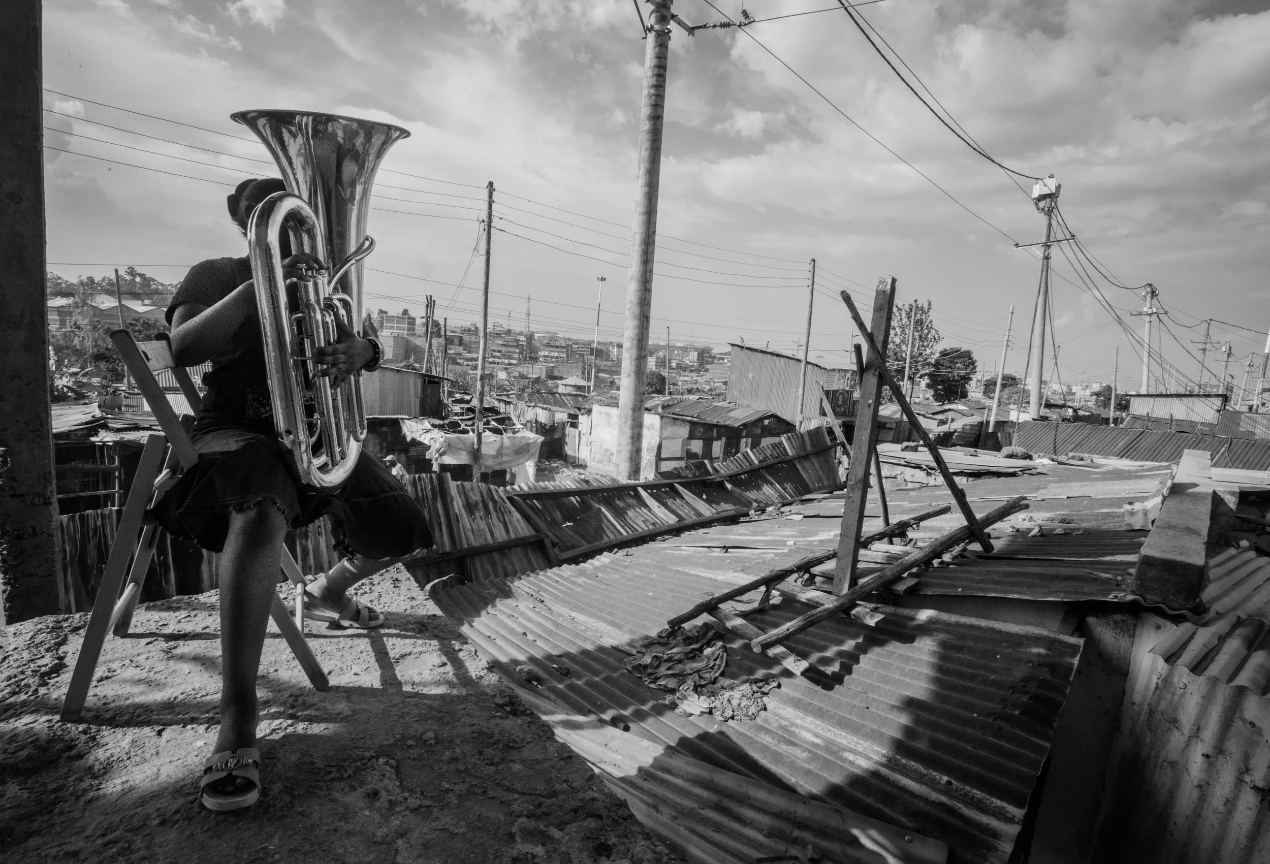 A person sitting on a plastic chair on a rooftop, playing a tuba, in an area with metal roofs and utility poles during daytime, black and white