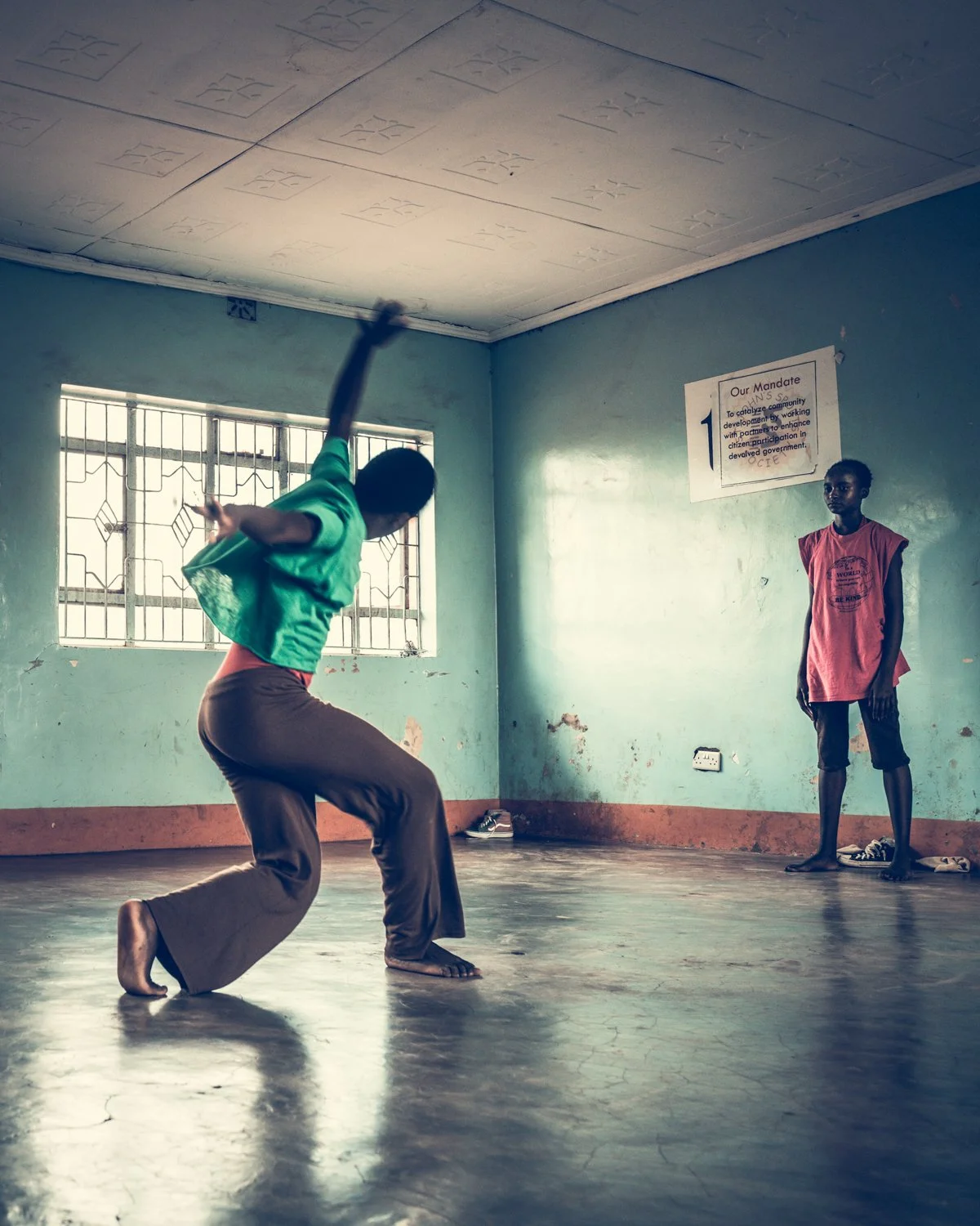 A woman in a green shirt and brown pants performing a dance move on a wooden floor while a girl in a pink shirt and black shorts watches in a room with pale green walls, barred windows, and a sign on the wall.