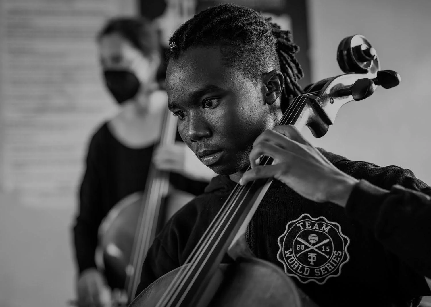 A young Black woman playing the cello in a music studio, wearing a hoodie with a 'Team World Series' logo, with another woman in the background wearing a mask.