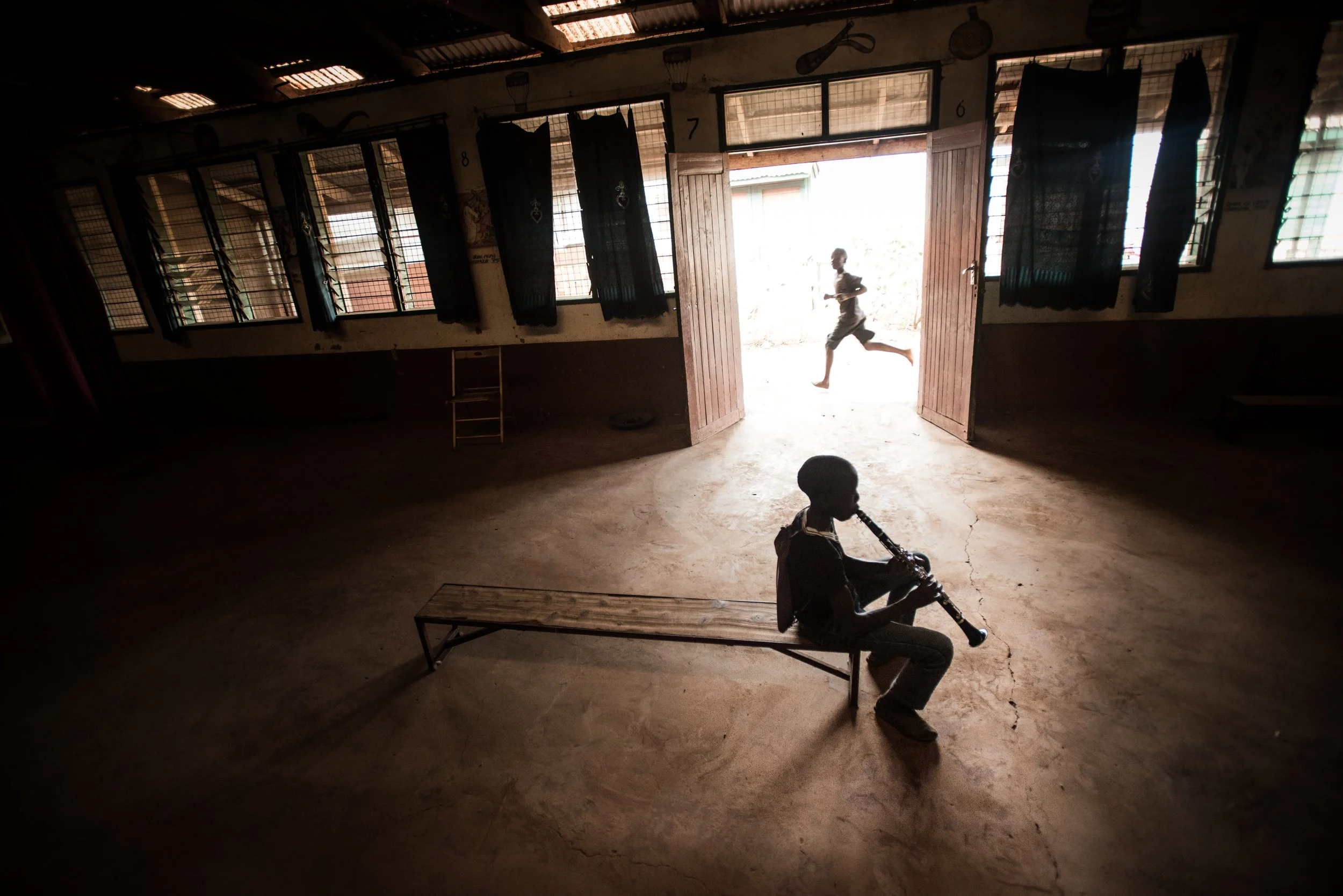 Silhouetted children in a dimly lit room with open doors and windows, one sitting on a bench playing a wind instrument, another running outside, with sunlight streaming in.