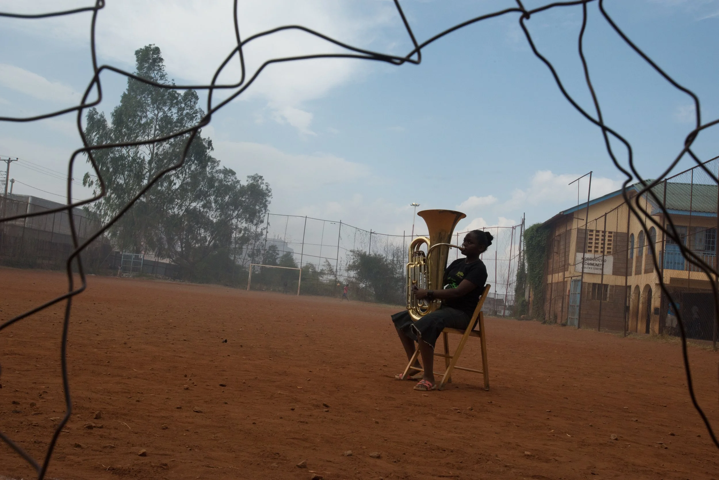 A woman sitting on a wooden chair on a dirt field, playing a tuba, seen through a wire fence, with trees and a building in the background.